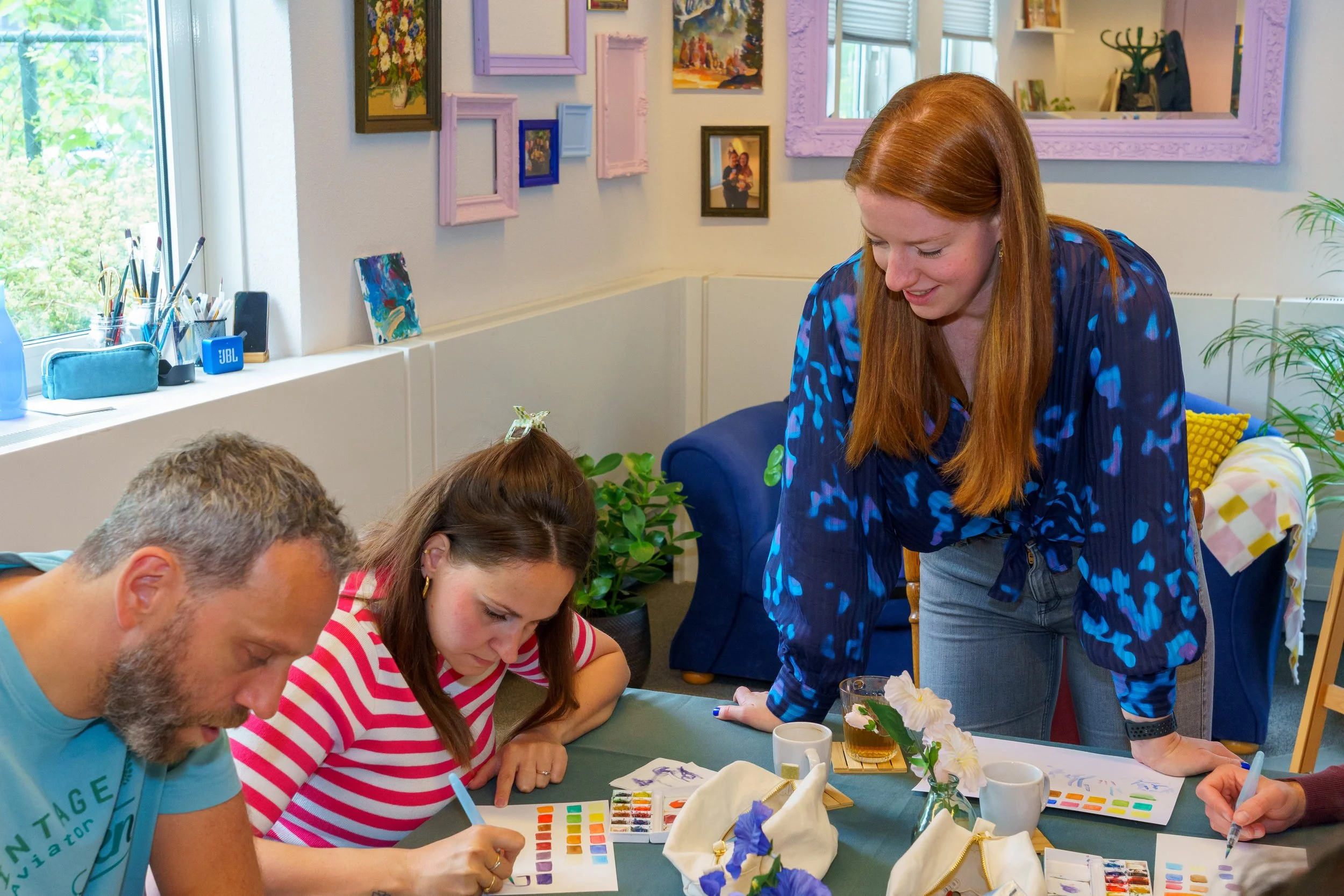 Vrouw begeleidt een groep mensen bij een aquarelschilderles in een kleurrijke, gezellige woonkamer.