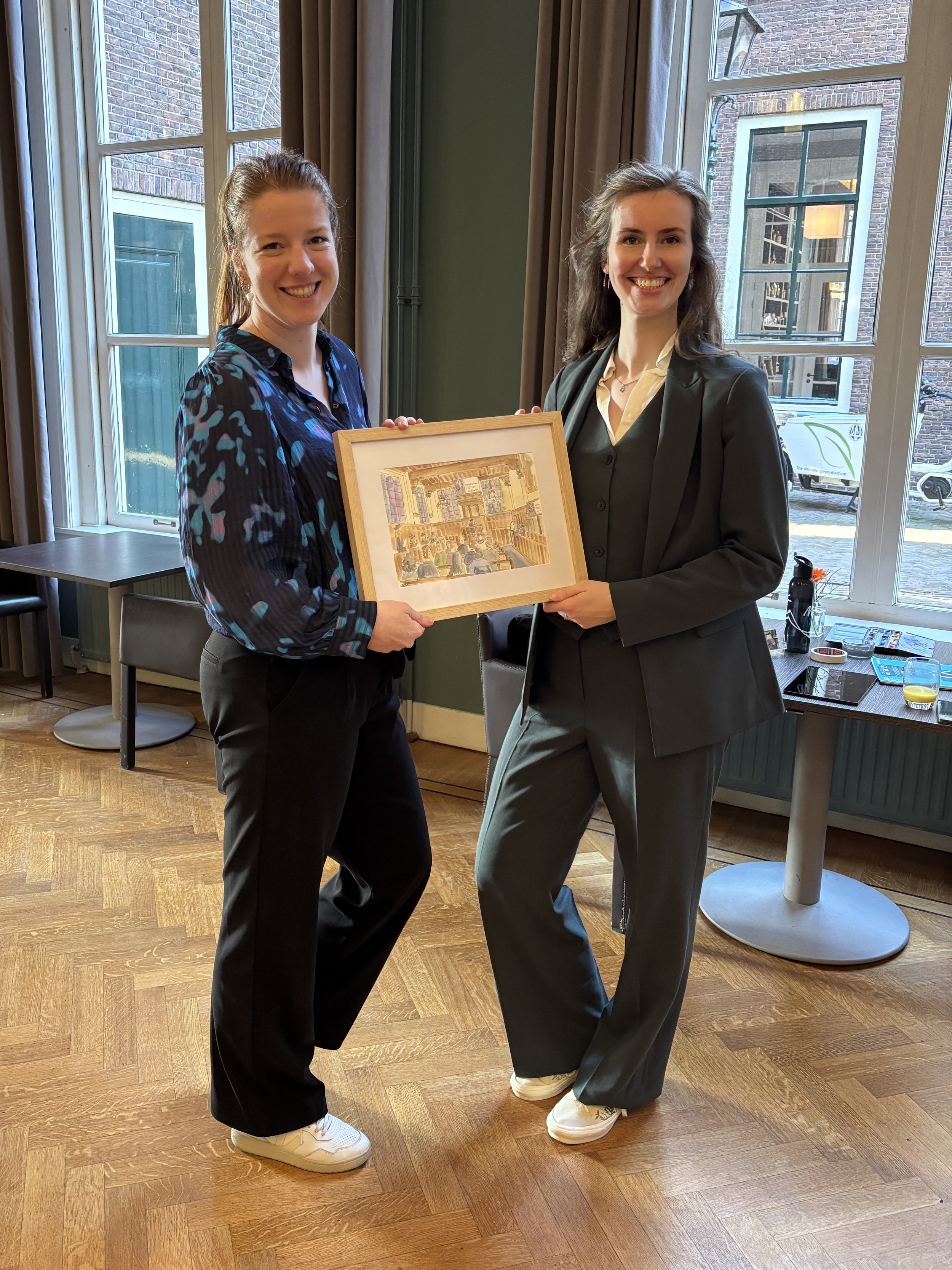 Twee vrouwen in een restaurant houden samen een ingelijste tekening vast, lachend naar de camera.