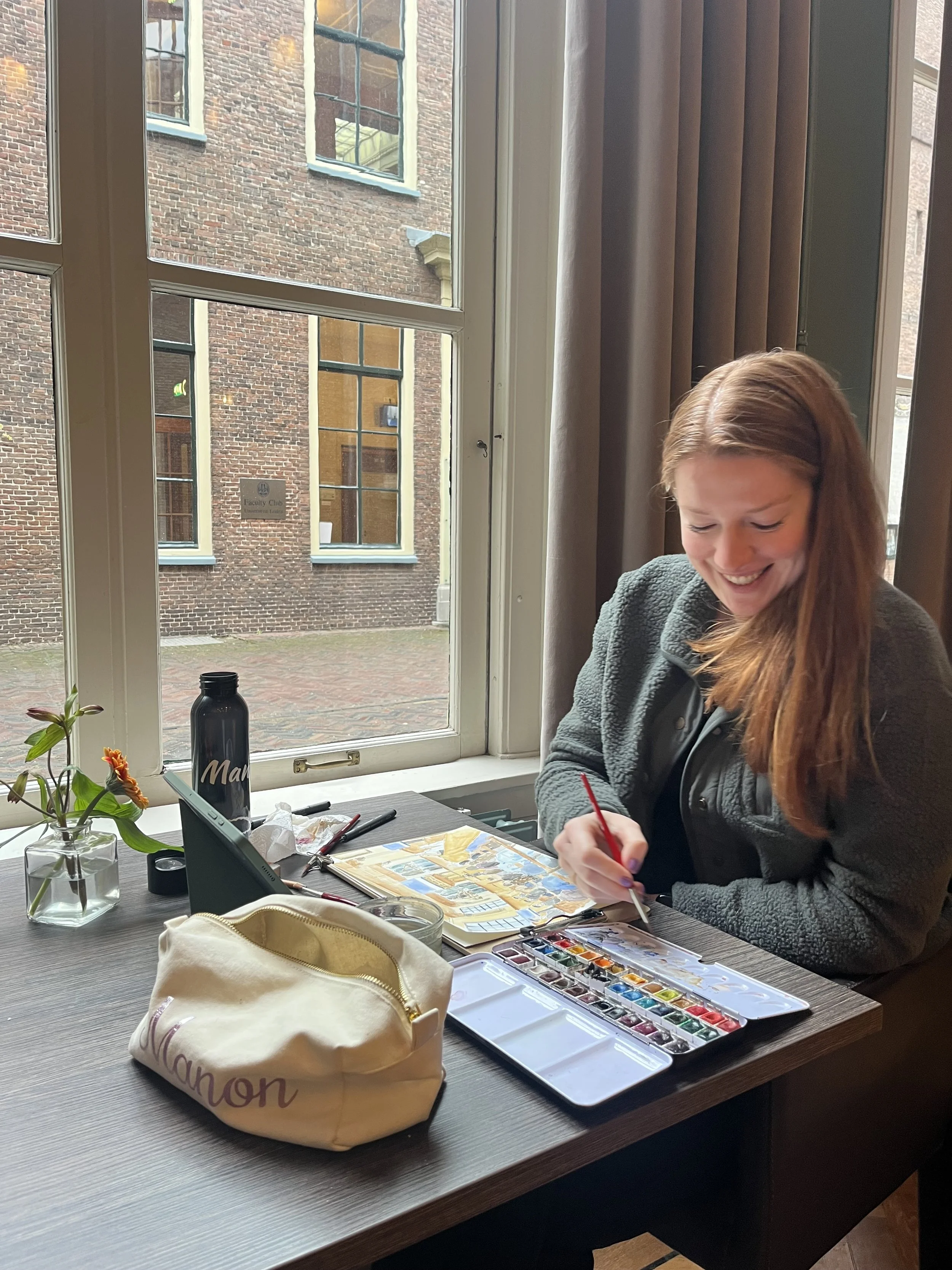 Vrouw met lang bruin haar die schildert met waterverf op een tafel met een open schilderdoos, naast een beker en een waterfles, bij een groot raam.