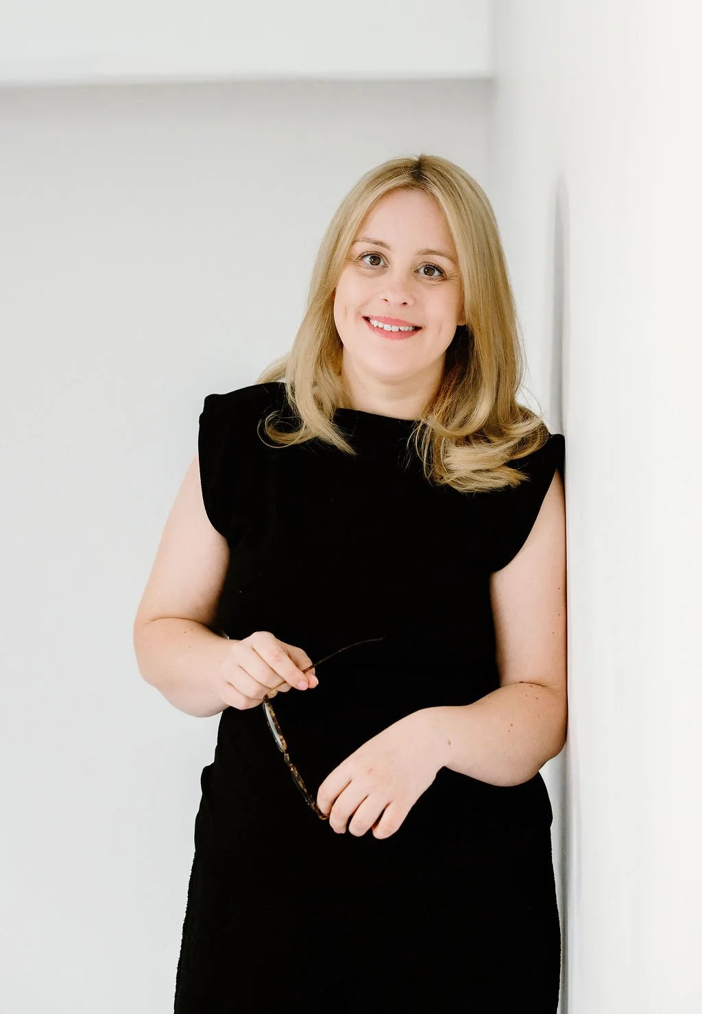 Portrait of a woman with blonde hair in a black dress, holding sunglasses, smiling, standing against a white wall.