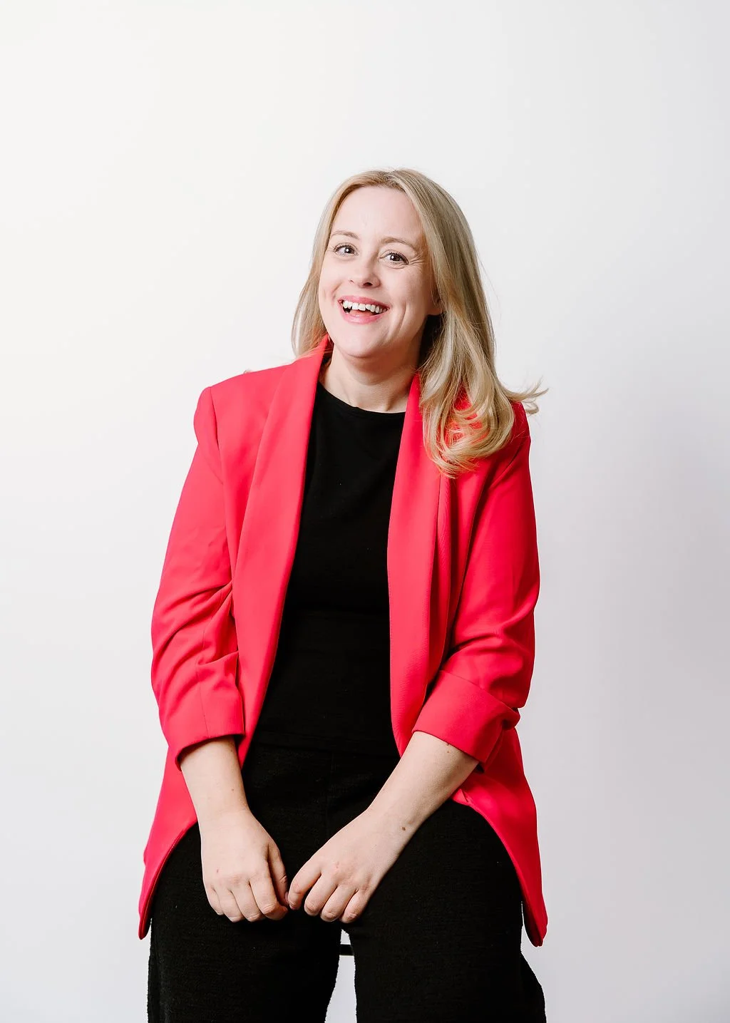 A woman with blonde hair smiling while sitting against a plain white background, wearing a red blazer over a black shirt.