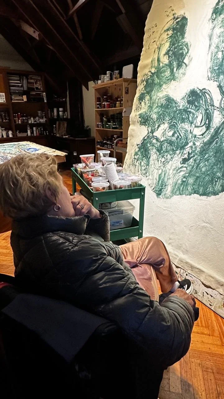 Mujer observando un mural en una galería de arte con una mesa de bocadillos y una estantería con frascos y artículos en el fondo.