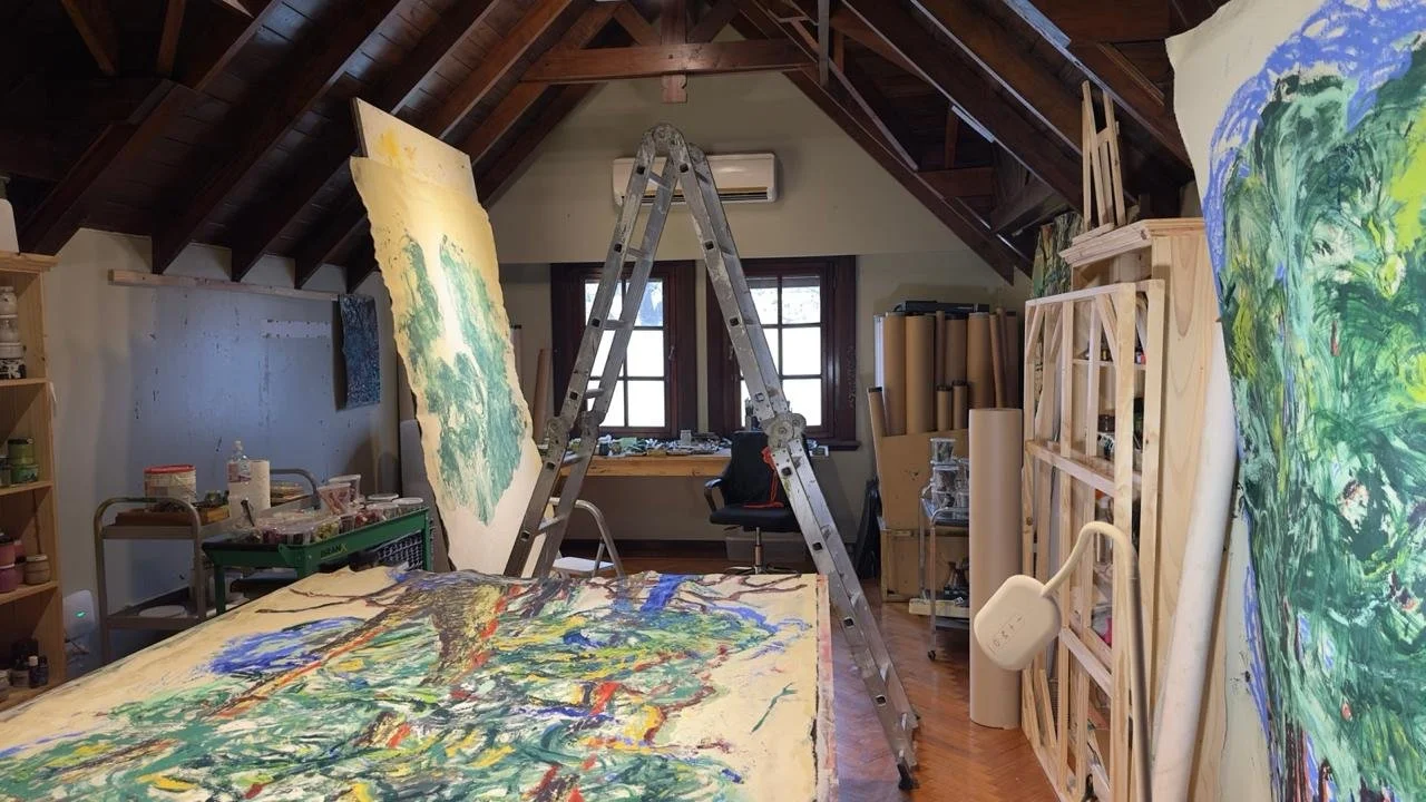 Estudio de arte con varias pinturas en proceso en las paredes, un caballete y una escalera de aluminio en el centro, ventanas al fondo y estanterías con materiales de pintura a los lados.