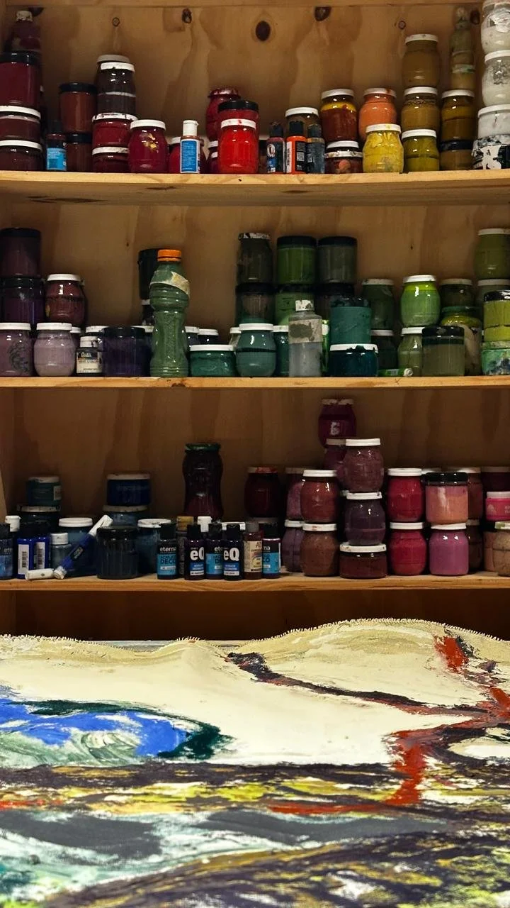 Estantes de madera con frascos de pinturas y tintas en diferentes colores, y parte de una obra de arte con pintura en la parte inferior.