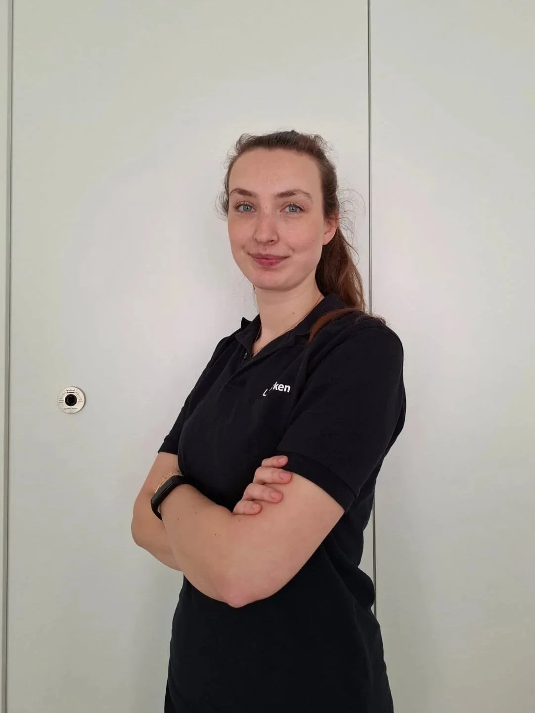 Junge Frau mit braunem Haar, gekleidet in einem schwarzen Polo-Shirt, steht vor einer weißen Wand mit geschlossenen Türen, Arme verschränkt, lächelt leicht.
