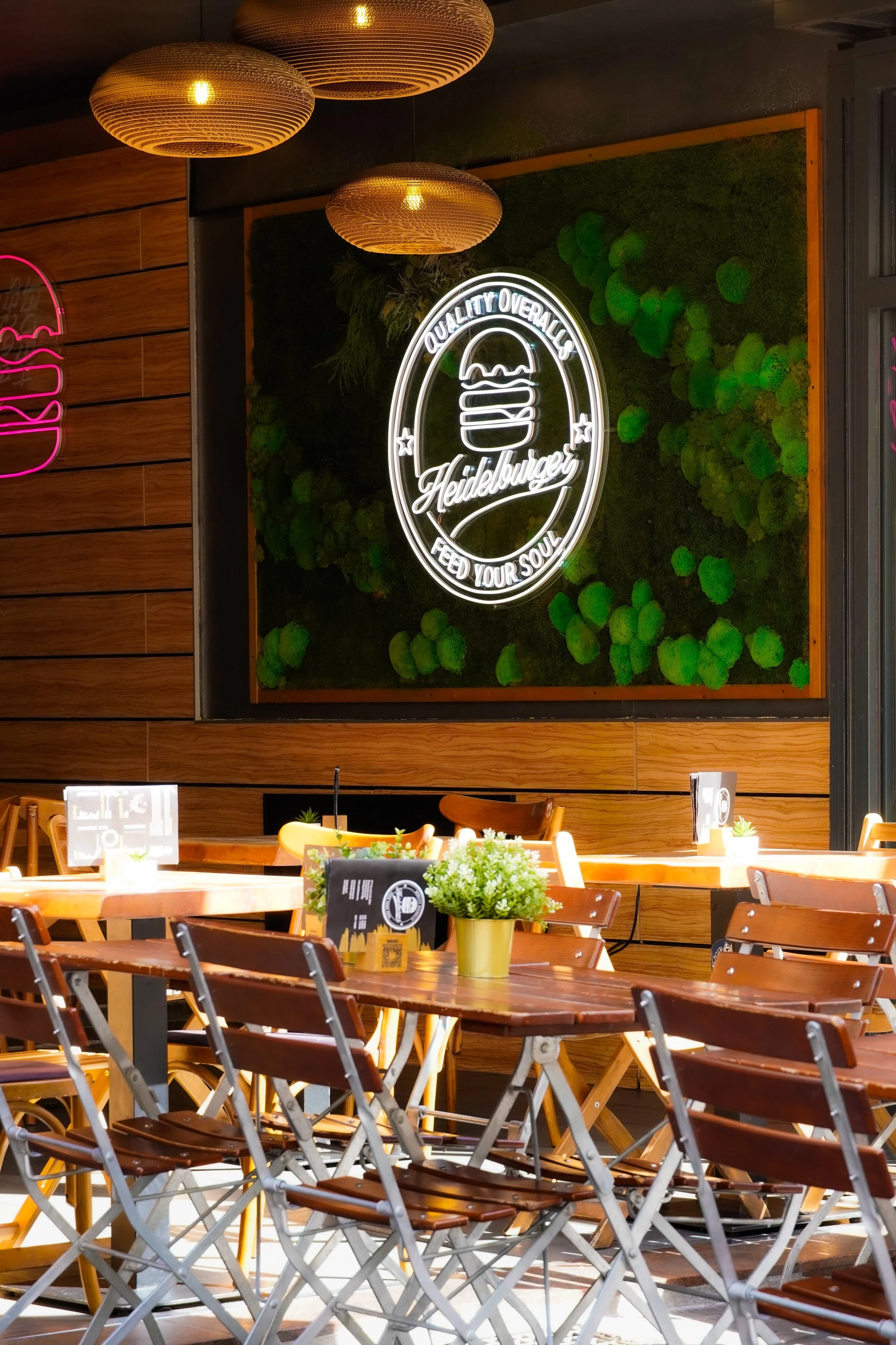 Ein gemütliches Café mit Holztischen und Stühlen, grünen Pflanzen an der Wand und einer neonbelichteten Tafel mit dem Schriftzug 'Heidelberger' sowie einem Kreis mit dem Text 'Qualität Overall Lidls' und 'Feed Your Soul'.
