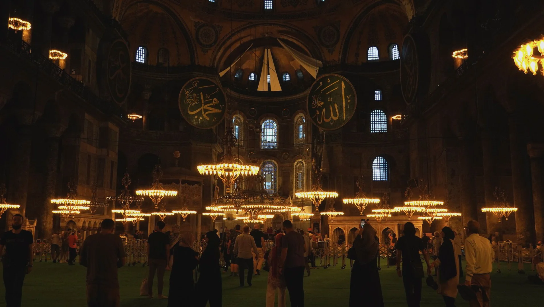 Innenraum einer Moschee mit großen Kronleuchtern, arabischer Kalligrafie an der Kuppel, vielen Menschen, die sich im Gebetsraum aufhalten, und buntem Licht durch die Fenster.
