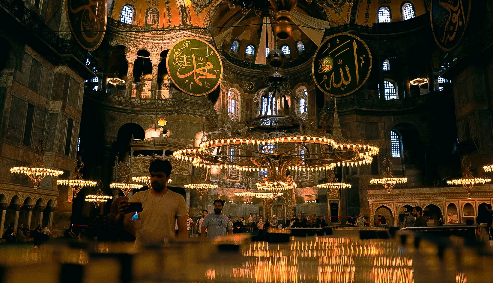 Innenraum einer Moschee mit großen goldenen Kronleuchtern, religiösen armenischen und islamischen Symbolen, mehreren Menschen, die die Moschee besuchen, und bunten Fenstern, die Licht ins Innere lassen.