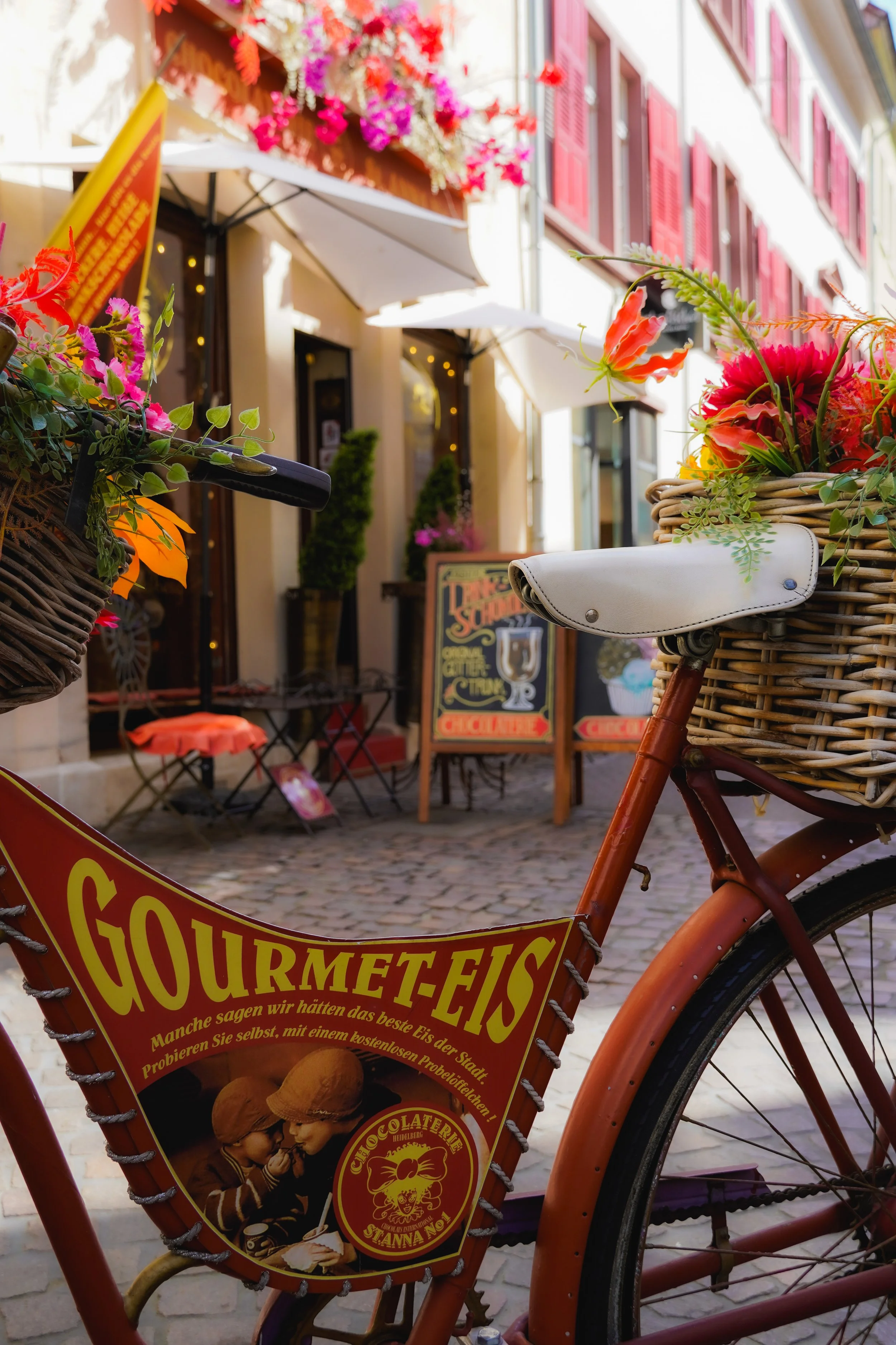 Ein Fahrrad vor einem Café mit Blumen und Schildern, in einem charmanten, lebhaften Stadtviertel.