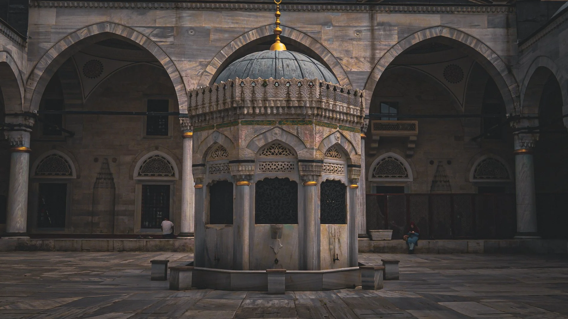 Ein Innenhof mit einem zentralen Brunnen in der Mitte, umlaufend Bögen und Säulen, typischer Marmorstil, im Hintergrund zwei Menschen, einer sitzt auf einer Bank, der andere steht.