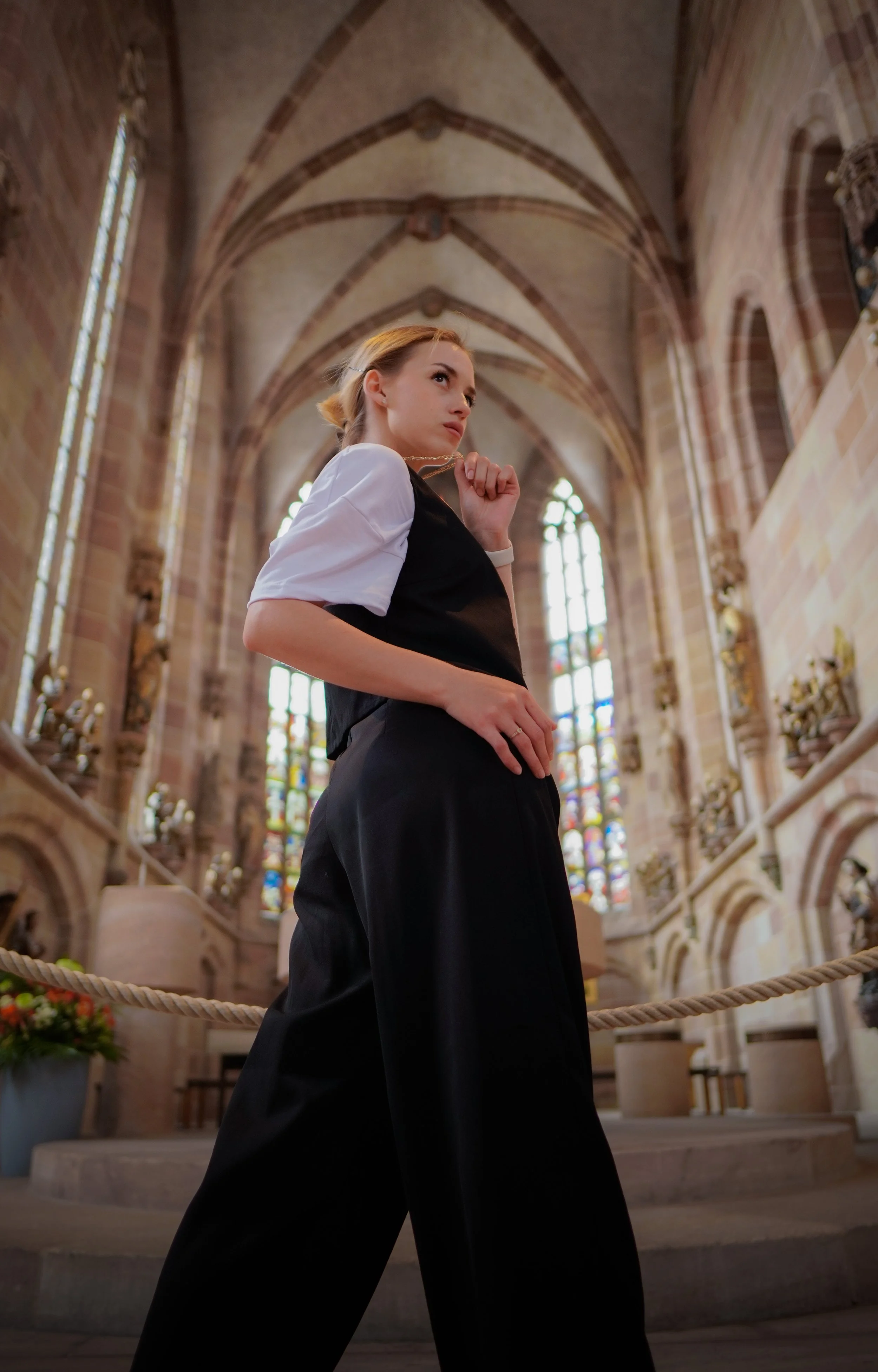 Junge Frau steht in einer gotischen Kirche mit hohen Fenstern und bunten Glasmalereien, trägt schwarze weite Hosen und ein weißes Hemd, wirkt nachdenklich.