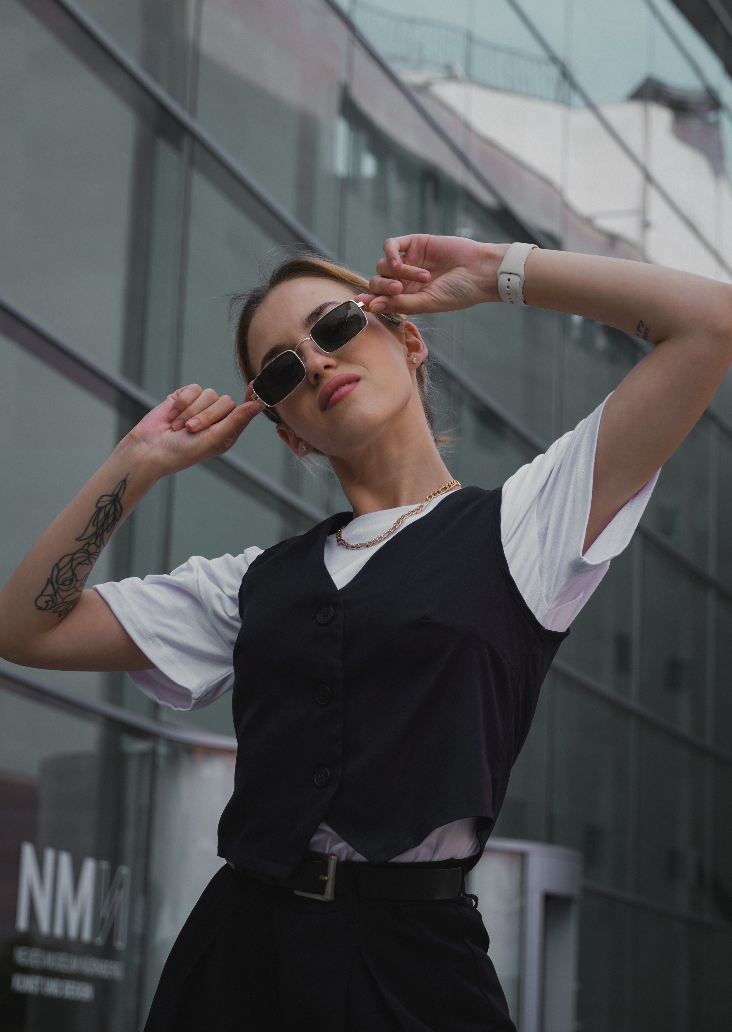 Eine Frau trägt schwarze Sonnenbrille, weiße T-Shirt und schwarzen Träger, posiert vor einem modernen Glasgebäude.