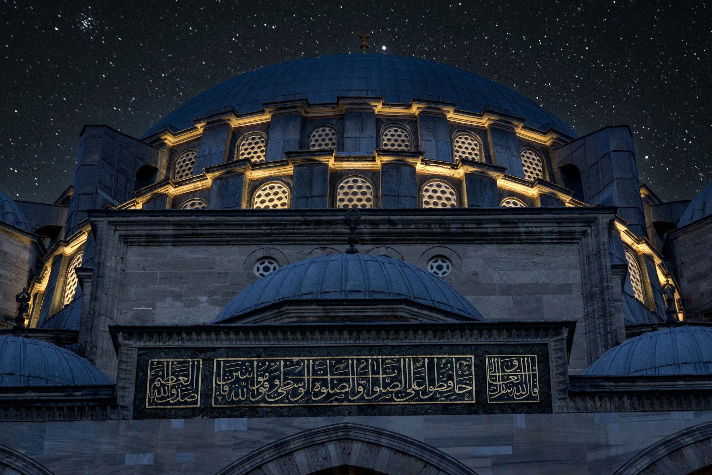 Nächtliche Aufnahme einer Moschee mit großen Kuppeln und einem Sternenhimmel im Hintergrund, die Kuppeln sind mit goldenen Linien beleuchtet, und es gibt eine schwarze Inschrift mit goldener Schrift auf einer schwarzen Platte am Eingang.