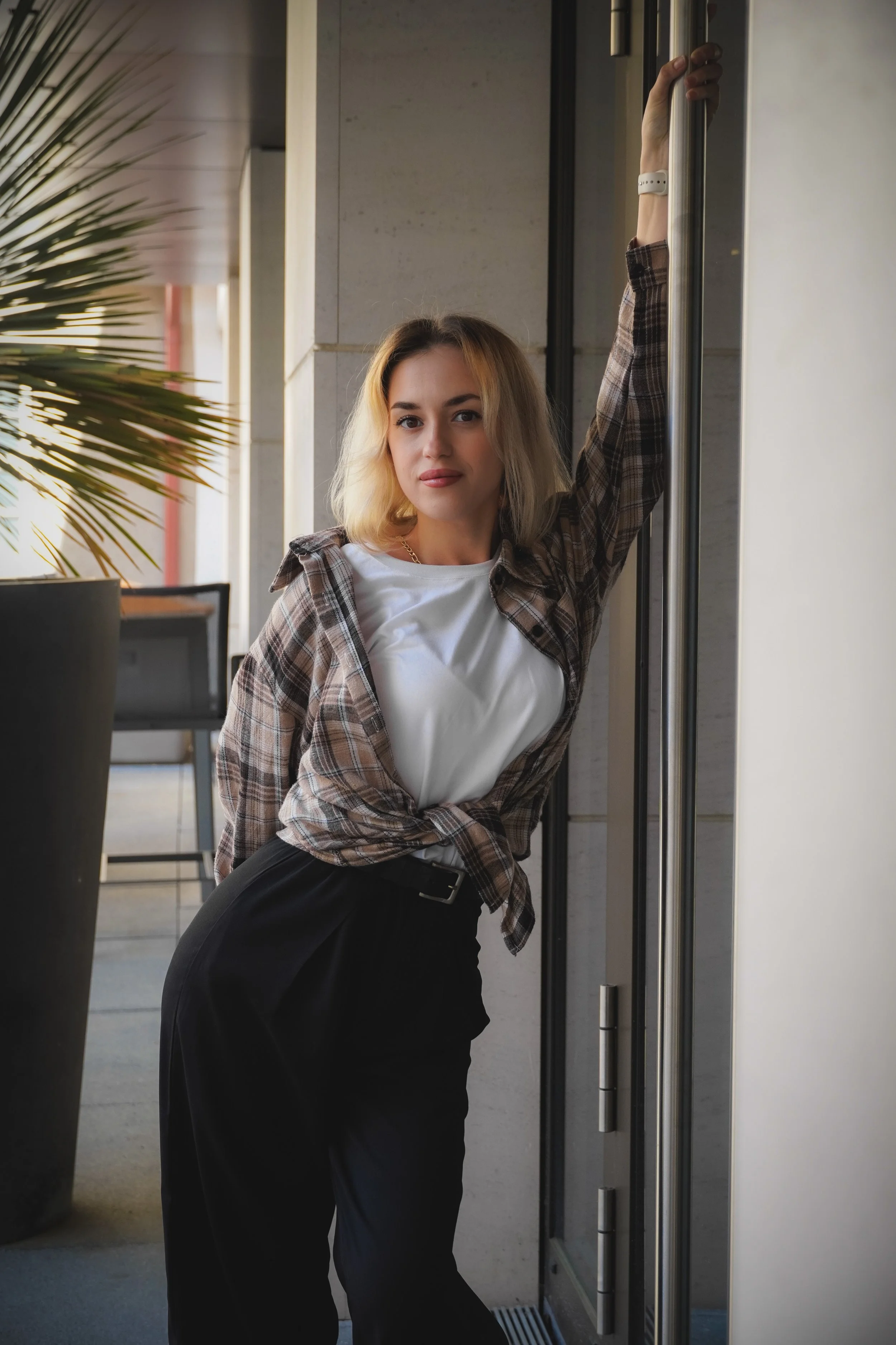 Junge Frau lehnt an einem Türrahmen, trägt eine kariertes Hemd, weiße T-Shirt und schwarze Hose, und schaut in die Kamera. Sie hat blondes, mittellanges Haar.