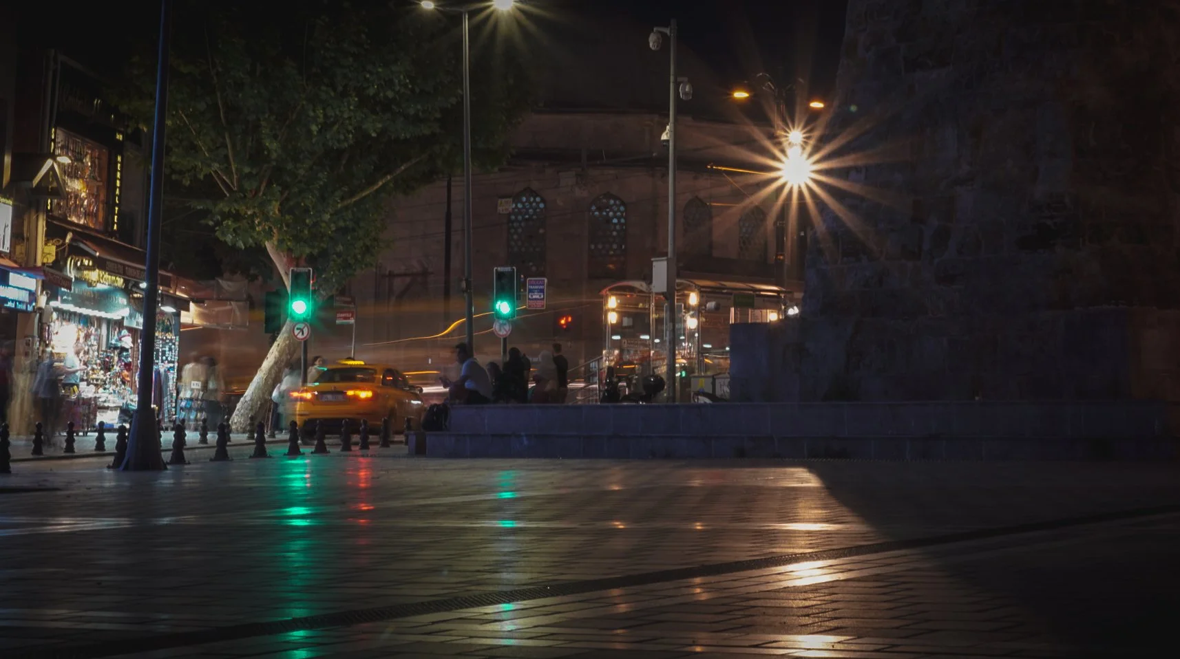 Straße bei Nacht mit Ampel, Taxis, Menschen, Straßenlaternen und im Hintergrund eine historische Gebäudewand.