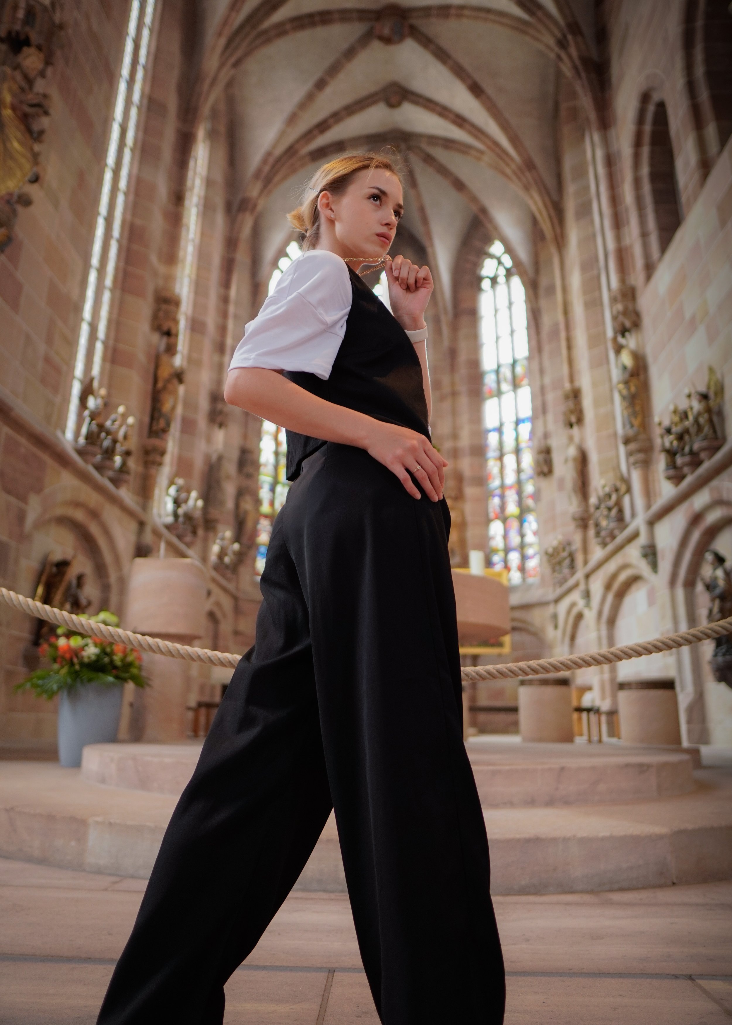 Junge Frau in schwarzen Hosen und weißem Hemd in einer Kirche mit hohen, bunten Fenstern und Steinwänden.