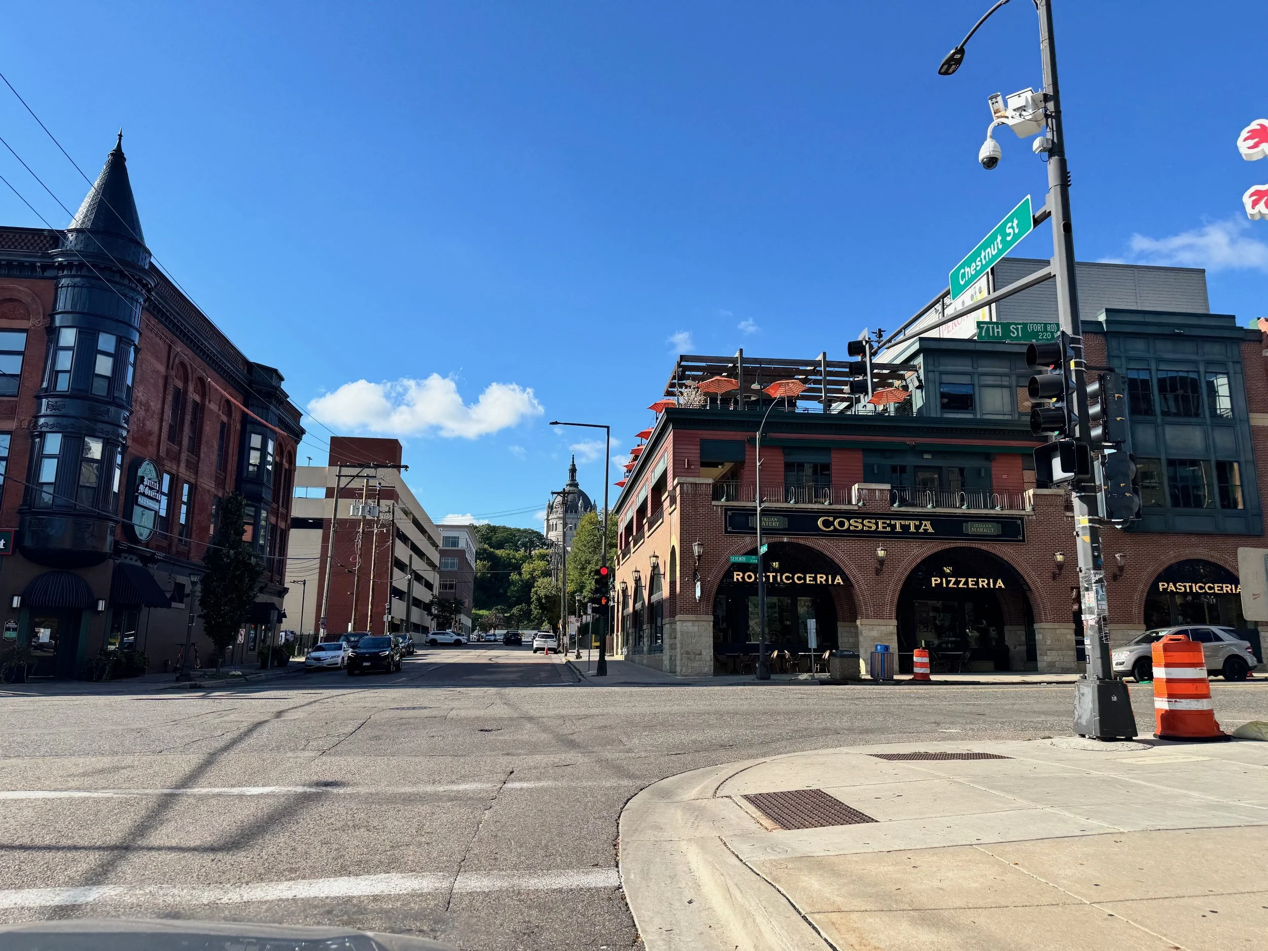 A city street corner with buildings, cars, and traffic lights under a blue sky with clouds. A prominent building on the right has signs for Cossetta, Rosticceria, Pizzeria, and Pasticceria. Street signs show Chestnut Street and 7th Street.