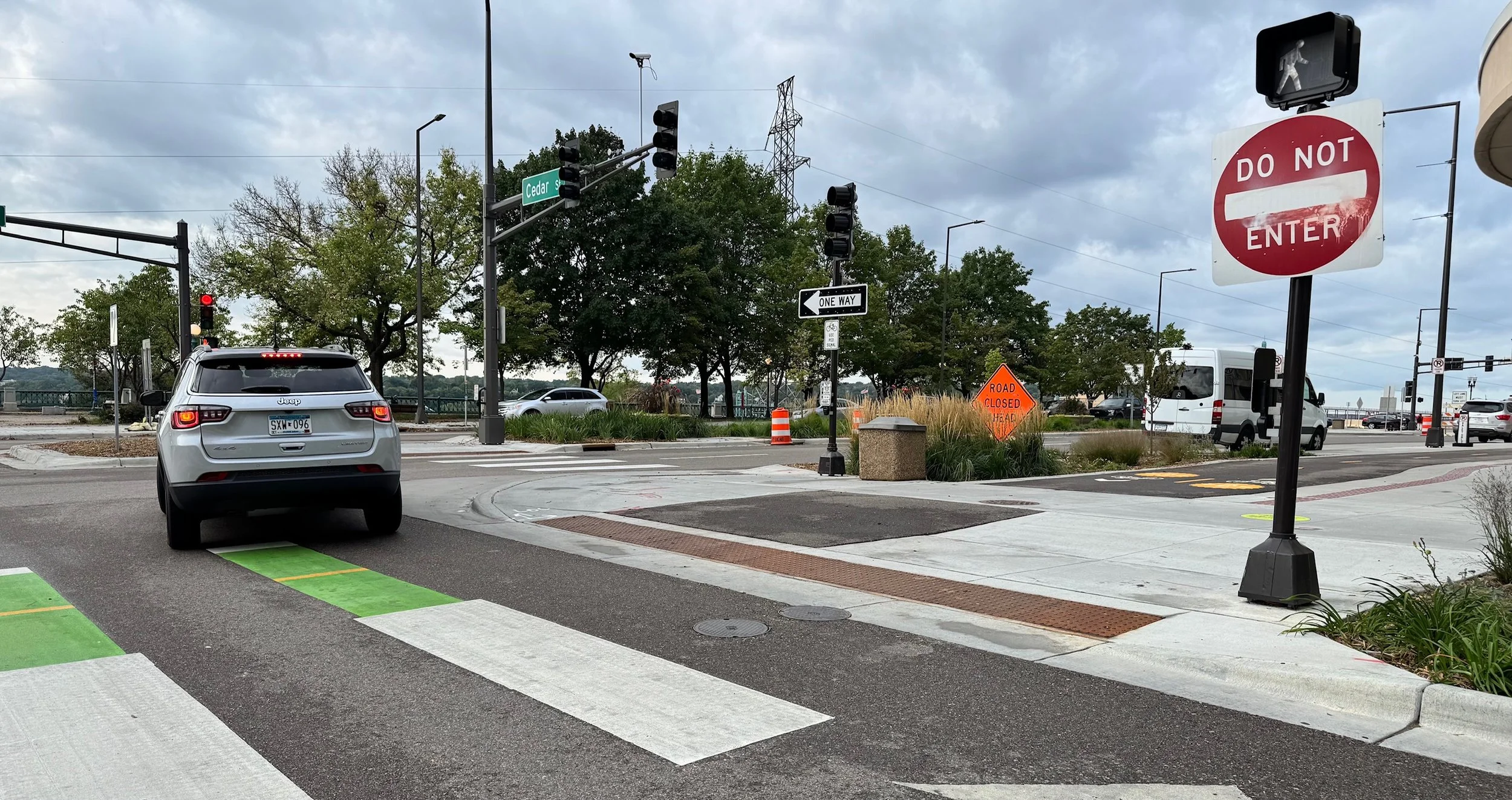 A white Jeep SUV stopped at a crosswalk with green markings, waiting to turn left at an intersection on a cloudy day. The intersection has traffic signals and street signs for Cedar Street. There is a red and white 'Do Not Enter' sign on a black pole, and a traffic light above the intersection. In the background, there is a construction sign indicating a closed road ahead, and a white van driving on the road. Trees are visible around the area.
