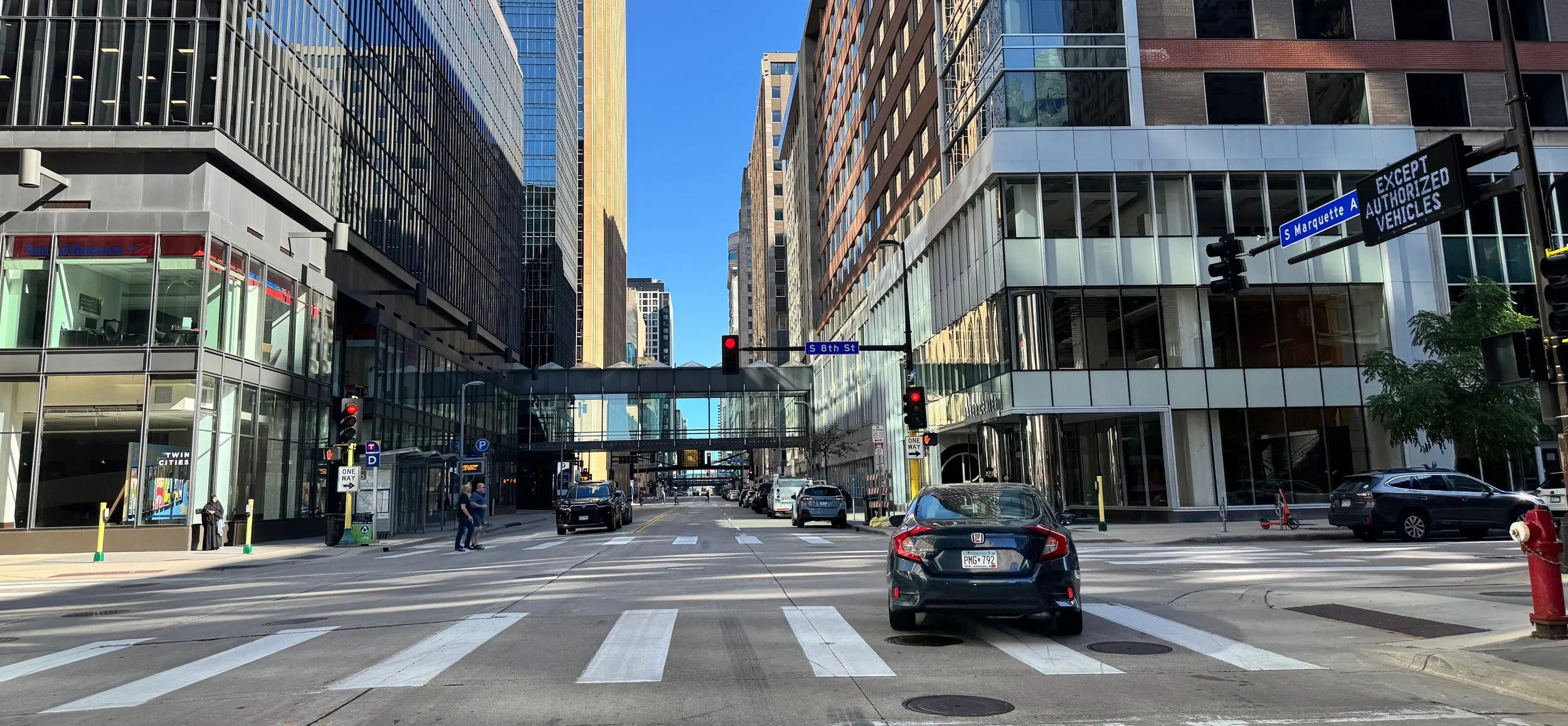 Minneapolis street view with tall modern buildings, crosswalks, traffic lights, vehicles, and pedestrians at an intersection during daytime.