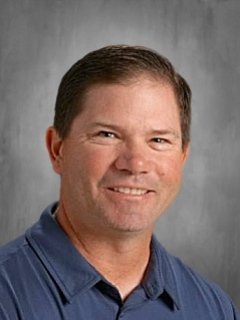 A smiling man with short, dark hair wearing a navy blue collared shirt, posed in front of a gray background.