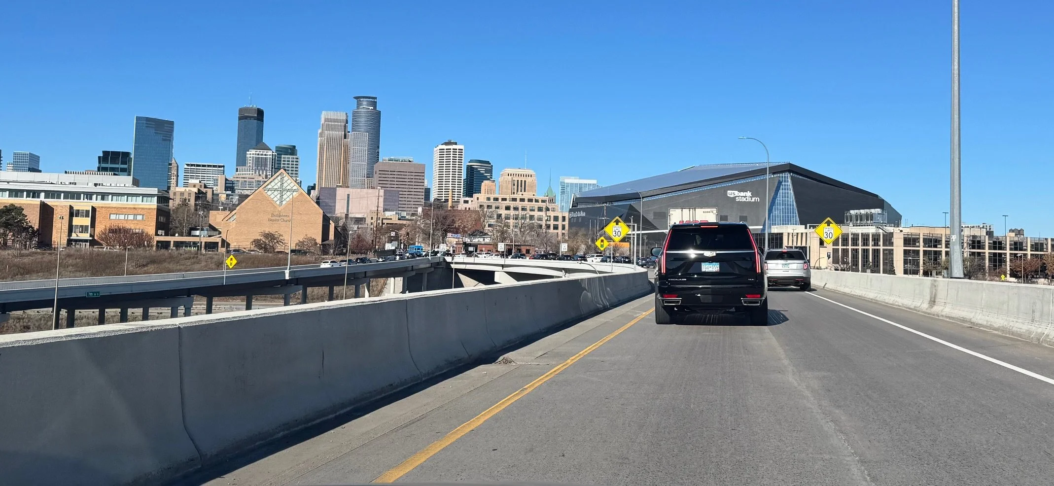 View of a city skyline with tall skyscrapers and U.S. Bank Stadium in the foreground, taken from a highway with cars traveling on it during a clear day.
