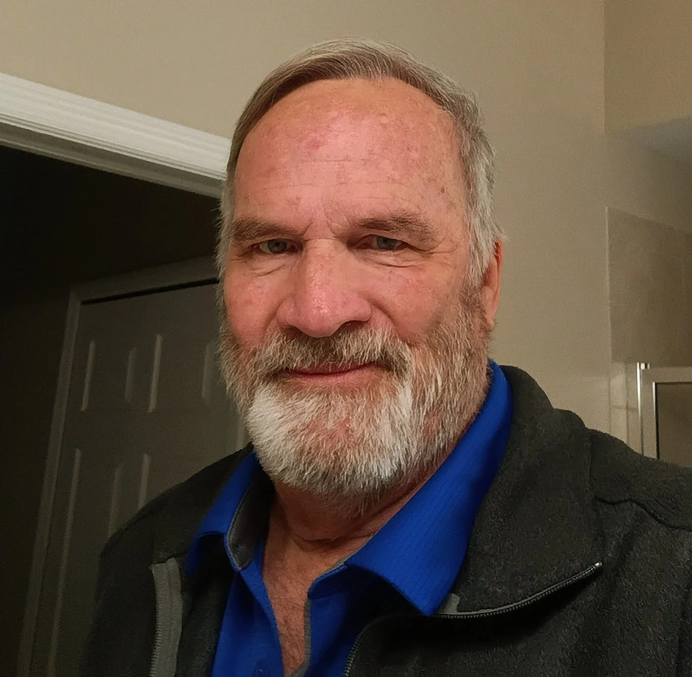 A middle-aged man with gray hair and a beard taking a selfie indoors, wearing a blue shirt and a dark jacket.