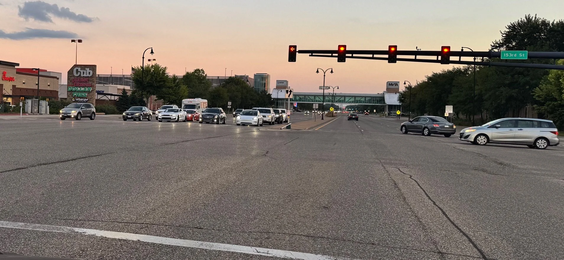 An intersection during sunset with traffic lights showing red signals, several cars waiting and moving, a pedestrian bridge, and buildings in the background including retail stores and street signs.