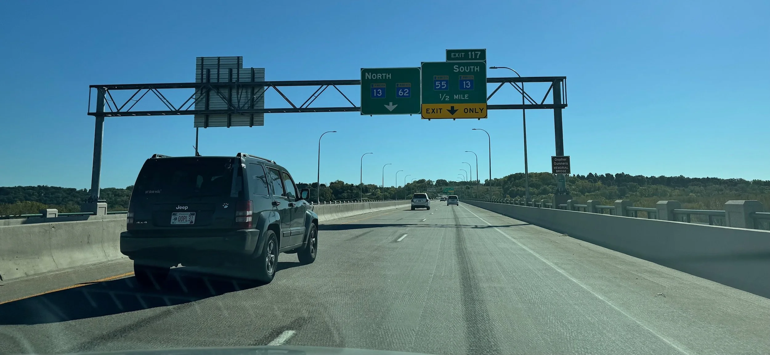 A highway view with green overhead signs indicating directions for North and South routes, exit information, and a few cars driving under bright blue sky, with a rural landscape in the background.
