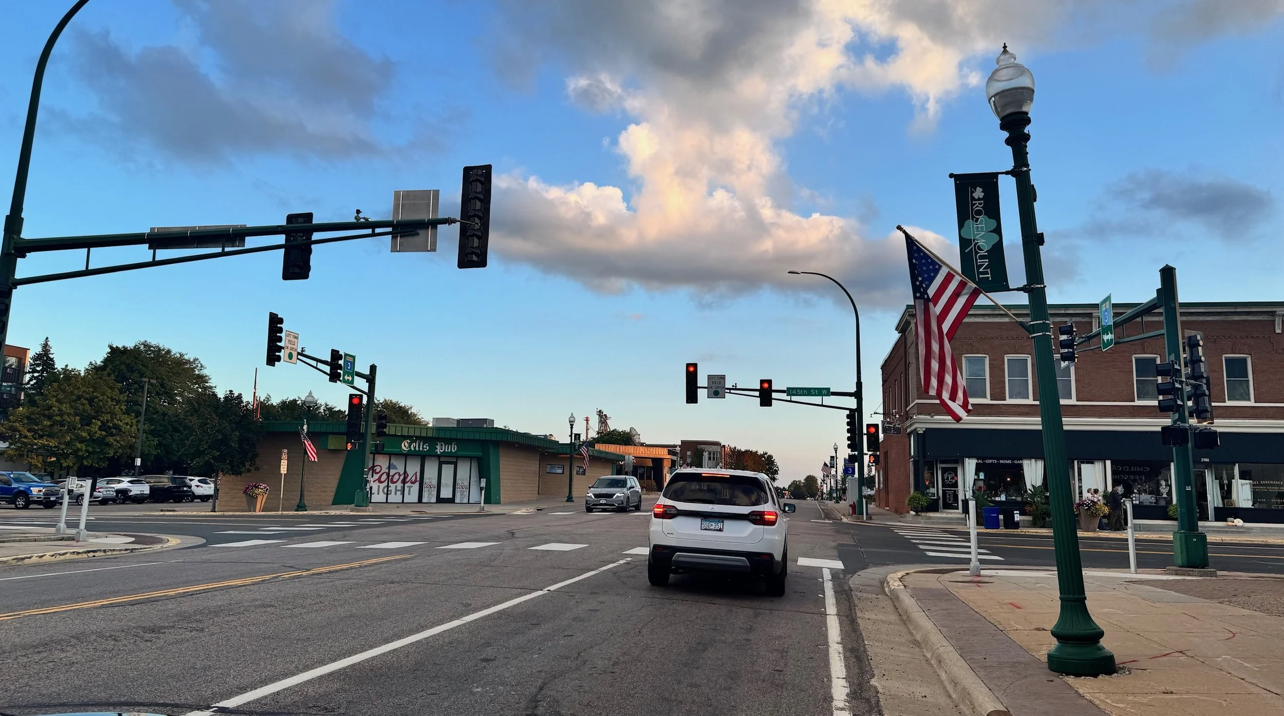 A street intersection with traffic lights, American flags, and a pub named 'Cats Pub' on the corner, under a partly cloudy sky.