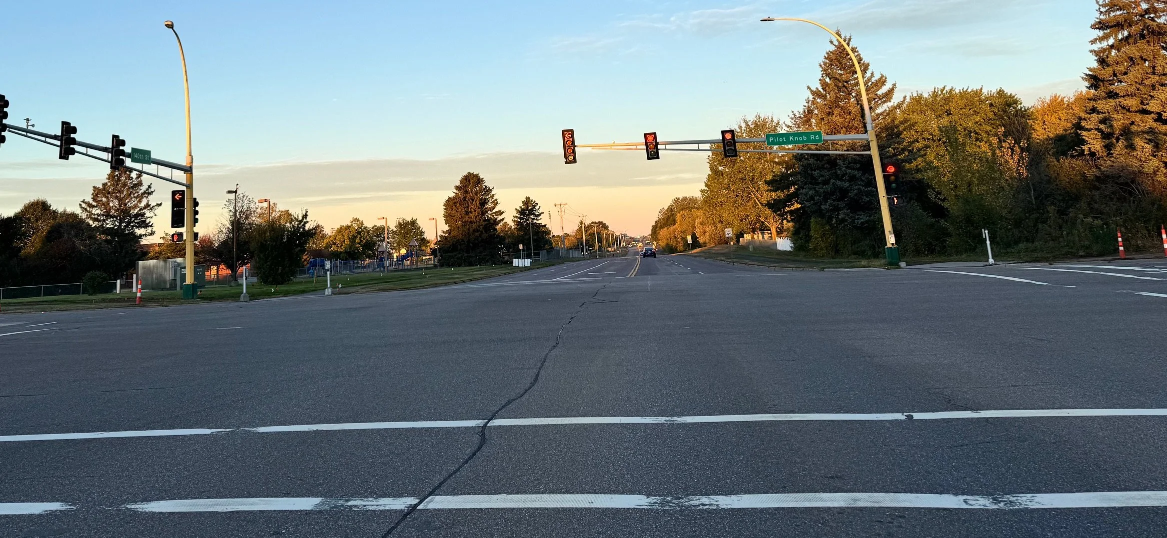 An intersection with traffic lights showing red, street signs for Pilot Knob Rd and 140th St, and trees with green and autumn-colored leaves on the sides.