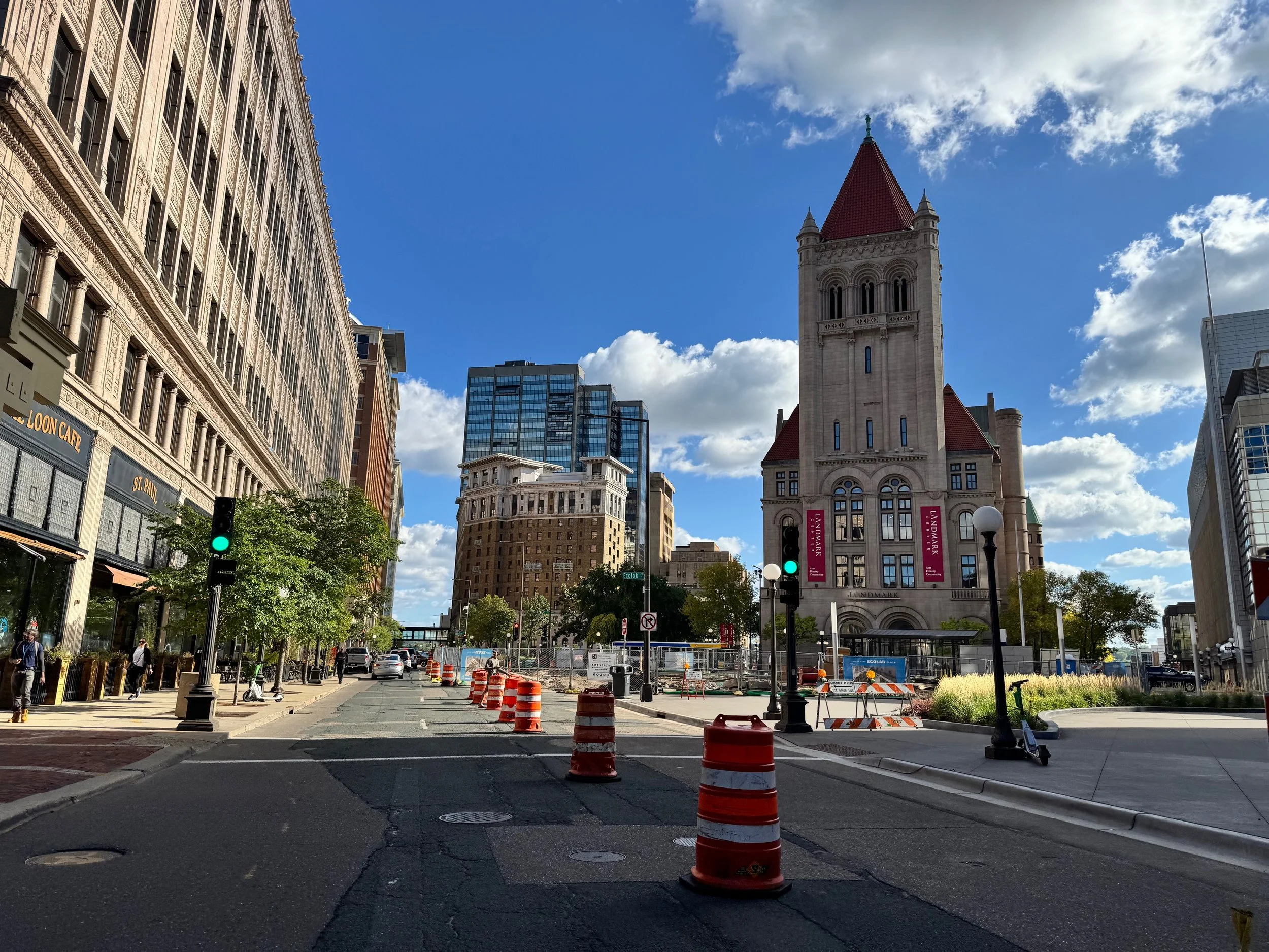 Urban street scene with construction cones, traffic lights, trees, and historic stone building with red roof and tall tower, surrounded by modern buildings under a partly cloudy sky.