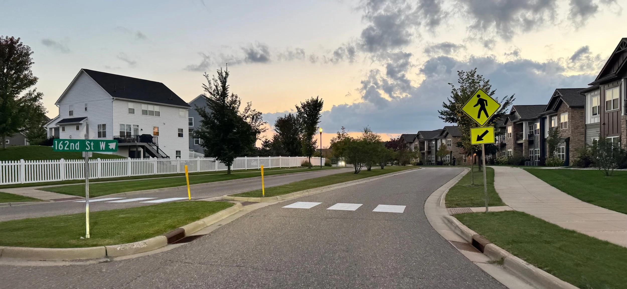 Residential street scene at sunset with houses, trees, street signs, crosswalk, sidewalk, and a white picket fence facing 162nd Street West.
