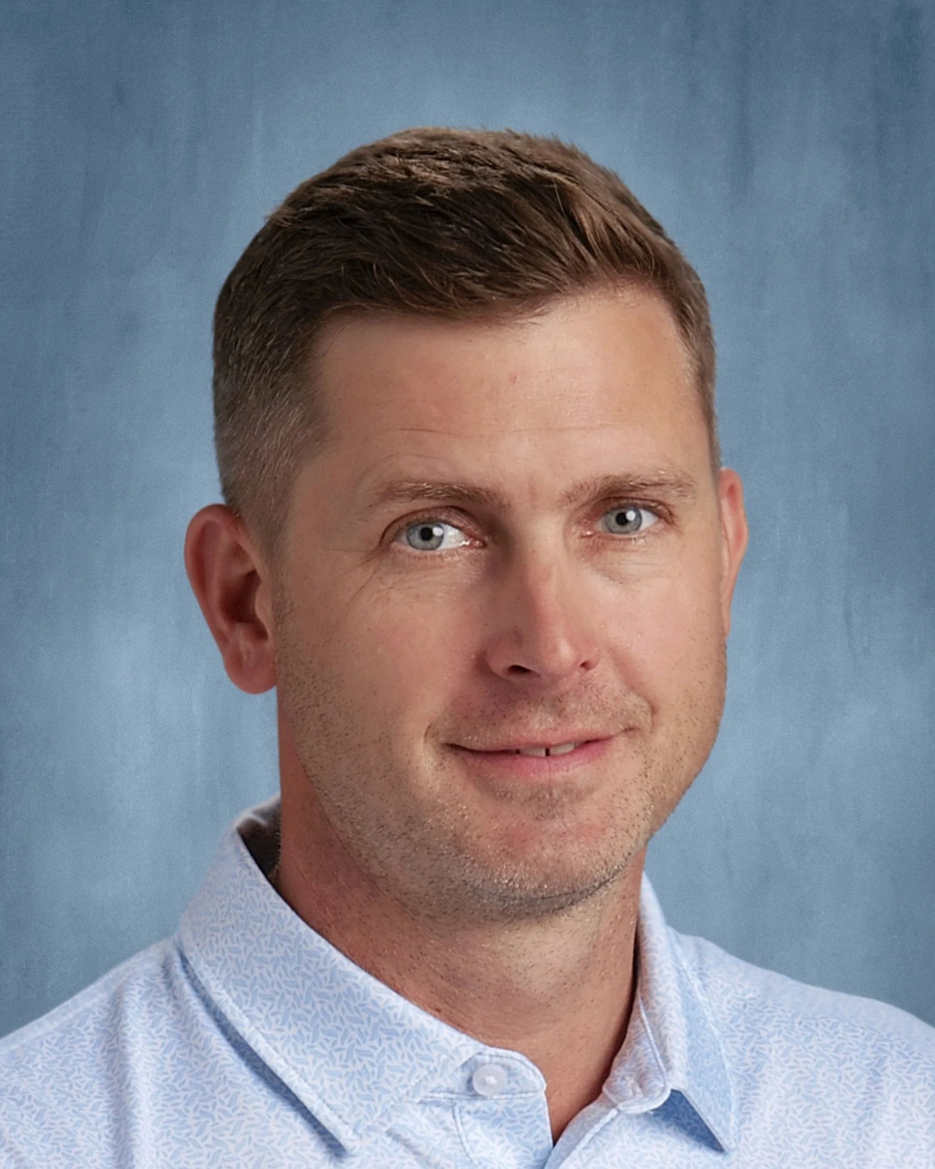 Portrait of Mr. Chris Scanlon with short brown hair, blue eyes, wearing a light blue patterned shirt, against a blue background.