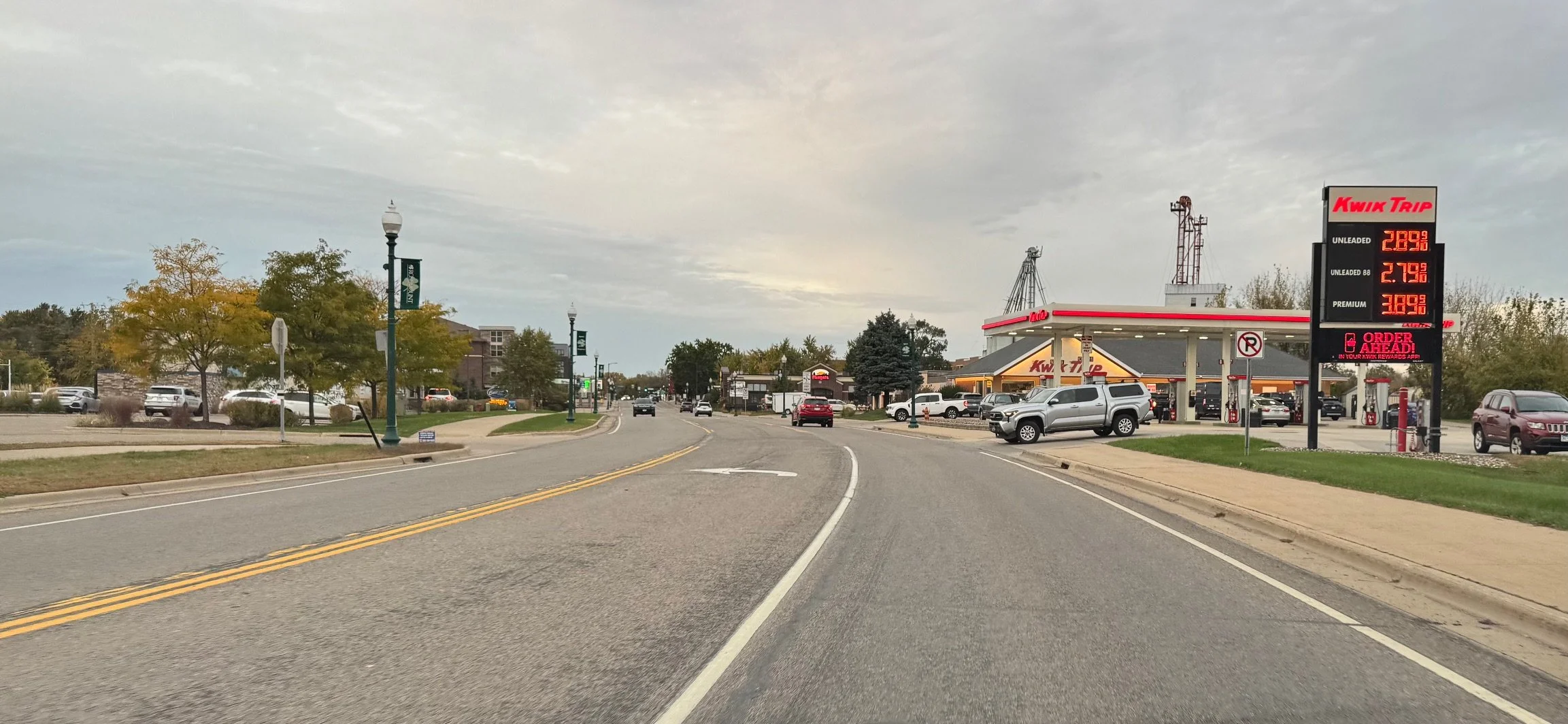 A street view in the evening with a Kwik Trip gas station on the right, displaying fuel prices on a digital sign. The street is lined with trees, lampposts, and cars parked along the sides, with some cars driving on the road. The sky is cloudy.