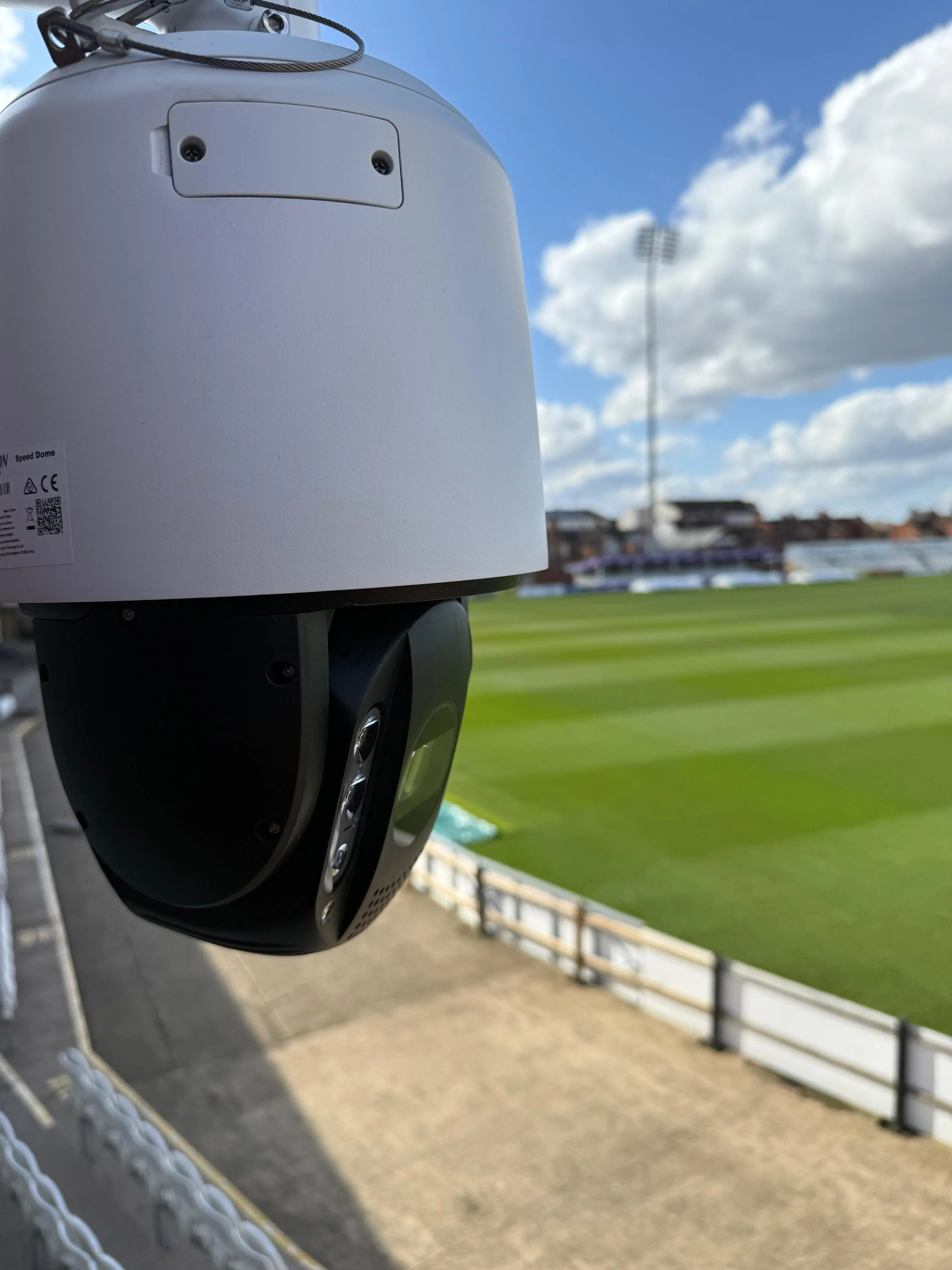 Close-up of a security camera mounted on a pole near a sports field with green grass, stadium lights, and cloudy sky in the background.