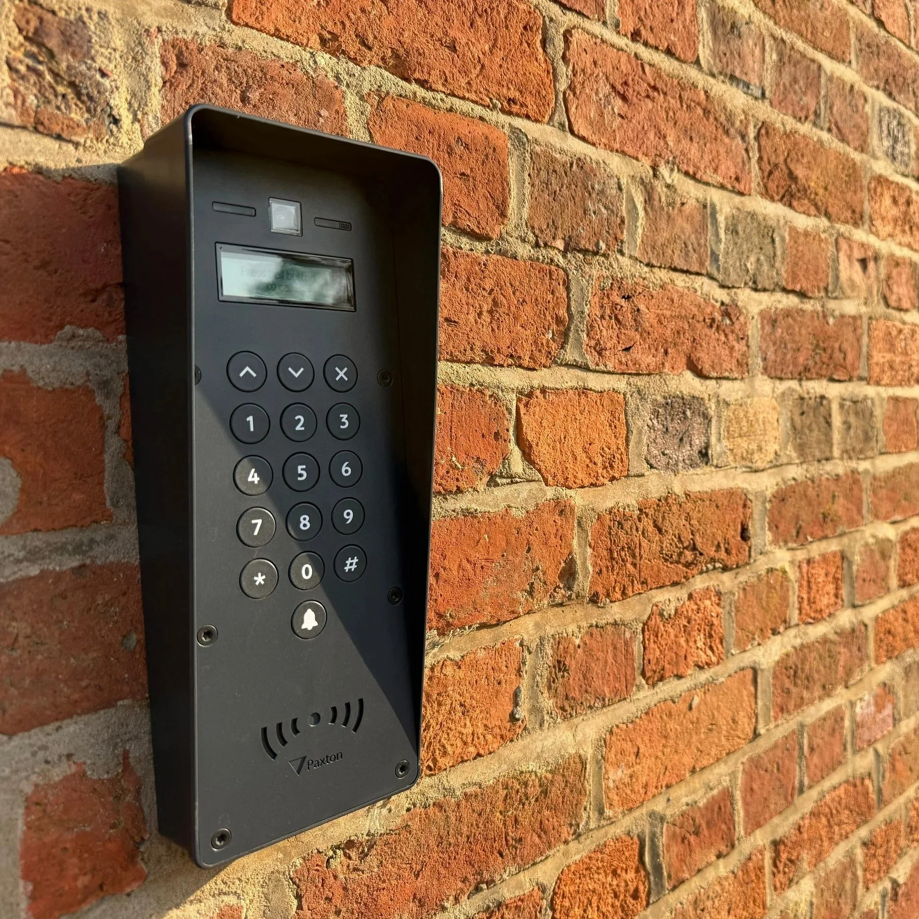 A black intercom panel with a small display, keypad, and bell button mounted on a red brick wall.