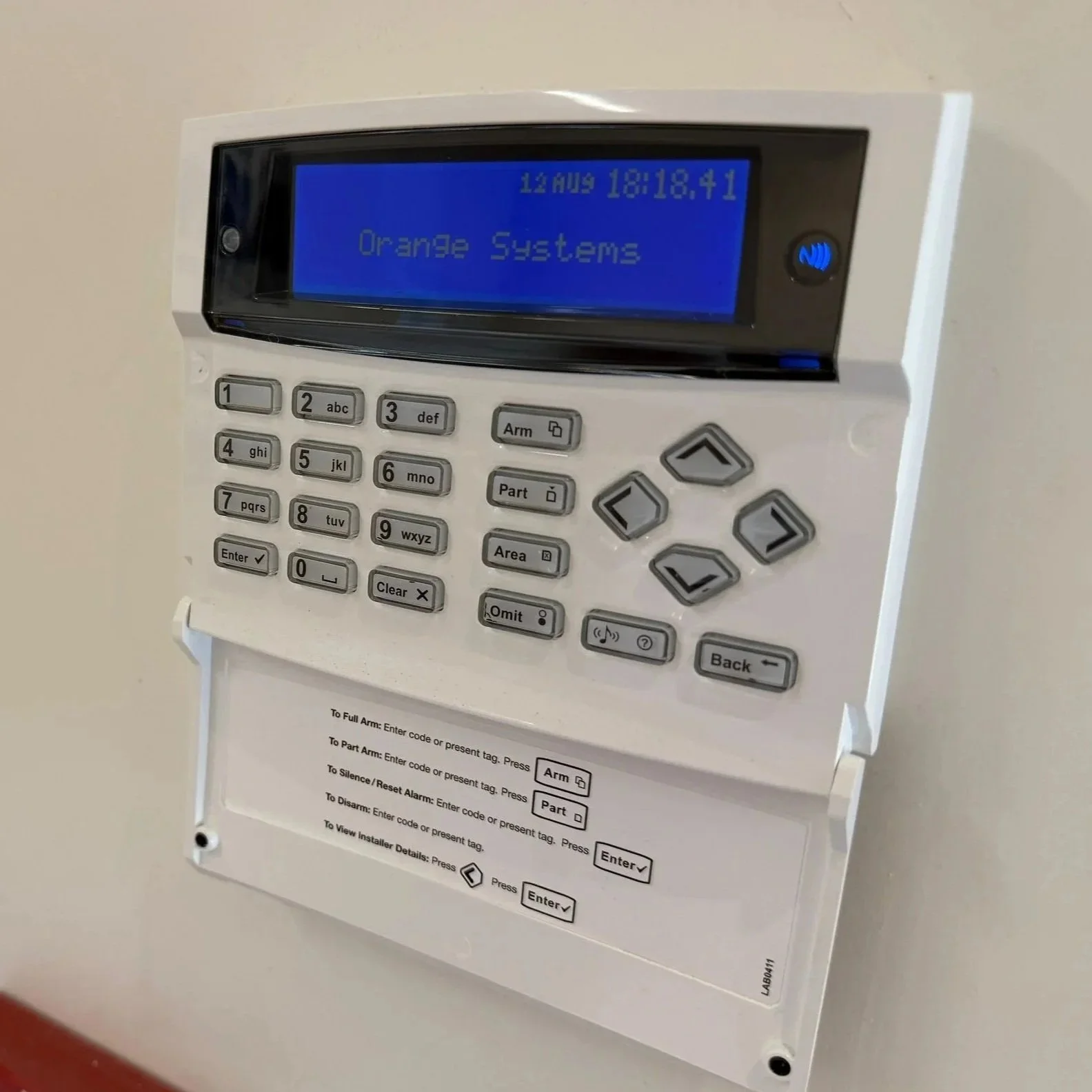 Security alarm control panel with a digital display showing the date, time, and 'Orange Systems' in blue background.