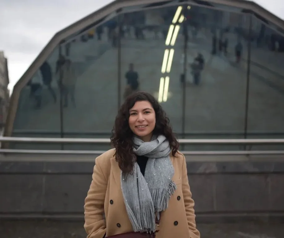 Une femme souriante devant un bâtiment aux façades en verre, avec des reflets de personnes et de lumières.