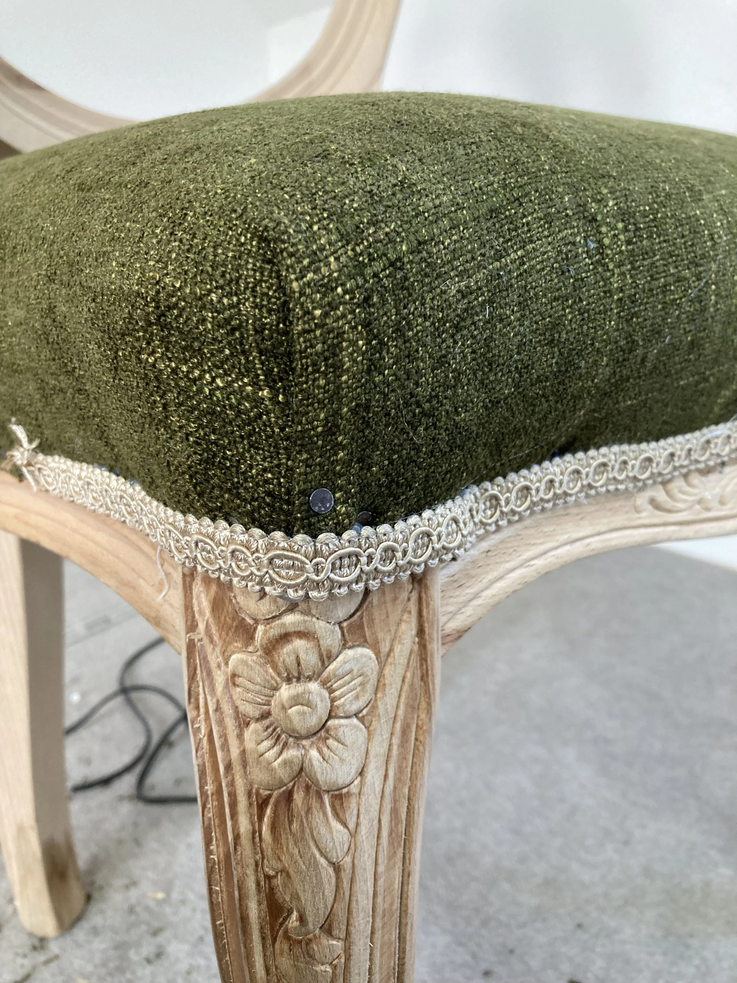 Close-up of a wooden table leg with detailed carved flower designs, a decorative trim, and green upholstered seat or cushion on top.