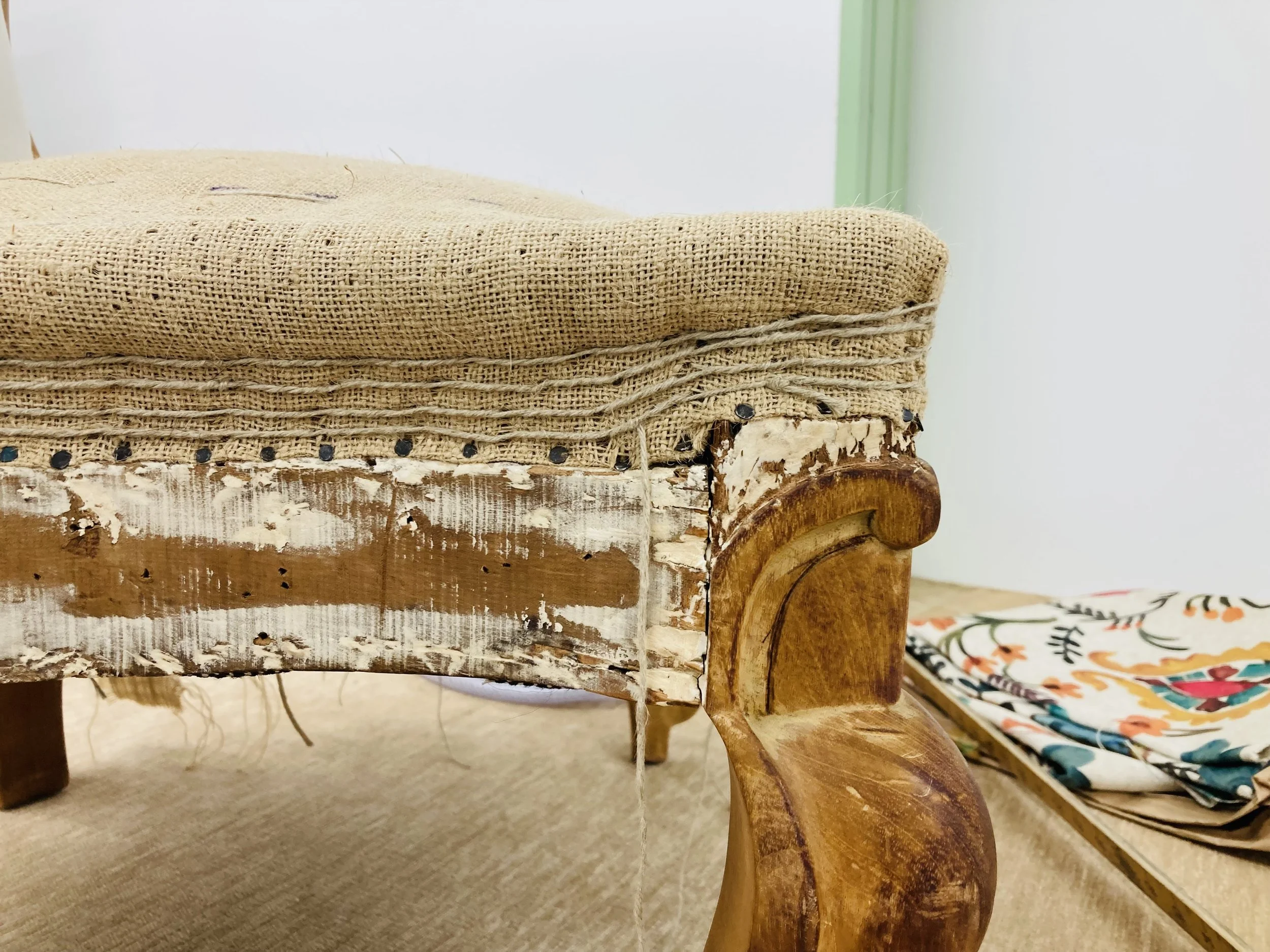 Close-up of a vintage wooden chair with a worn fabric cushion on top, placed next to a colorful floral-patterned fabric.
