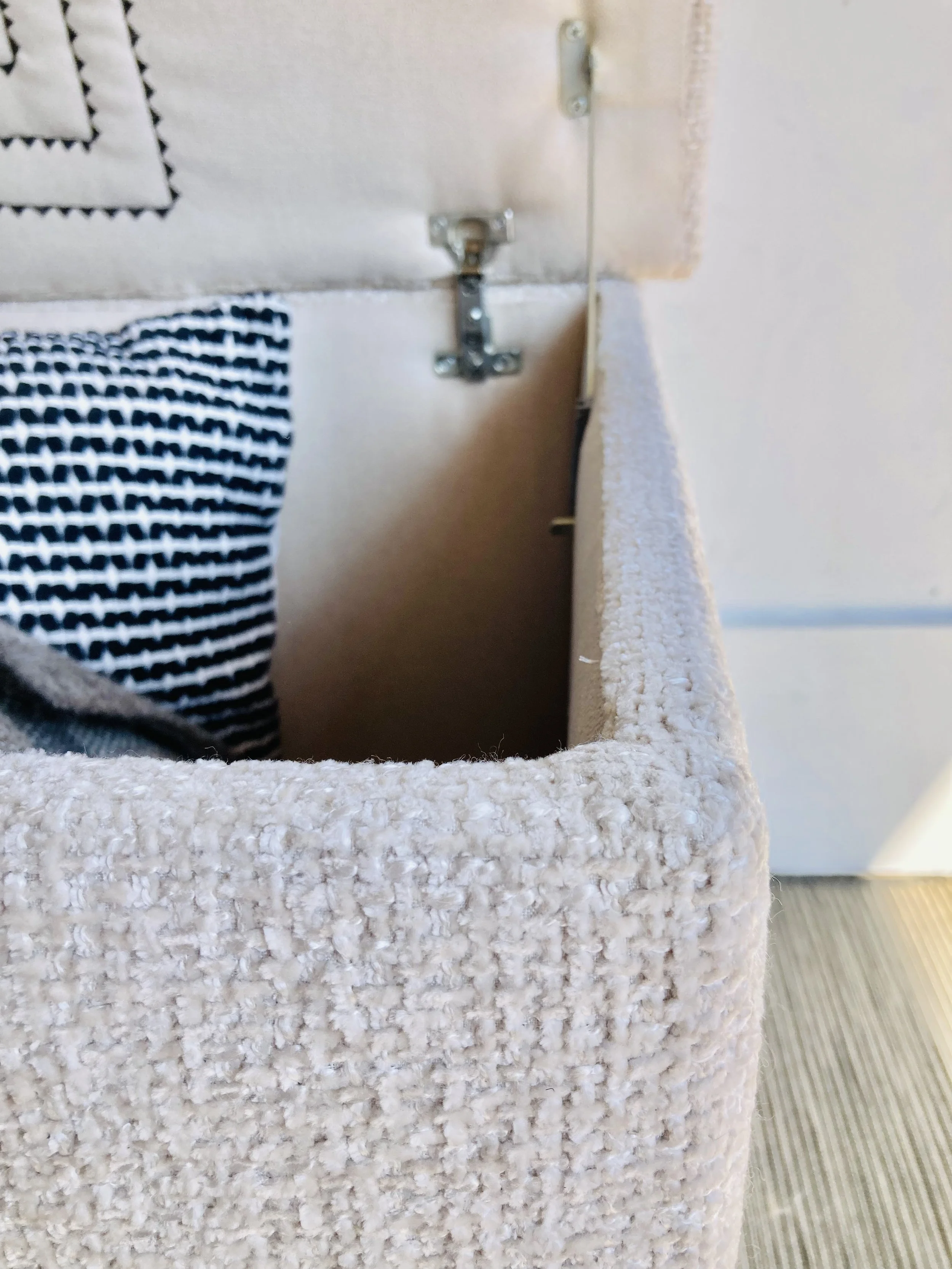 Close-up of a textured beige fabric box with a metal hinge, showing a partial view of a striped fabric inside and a beige wall in the background.