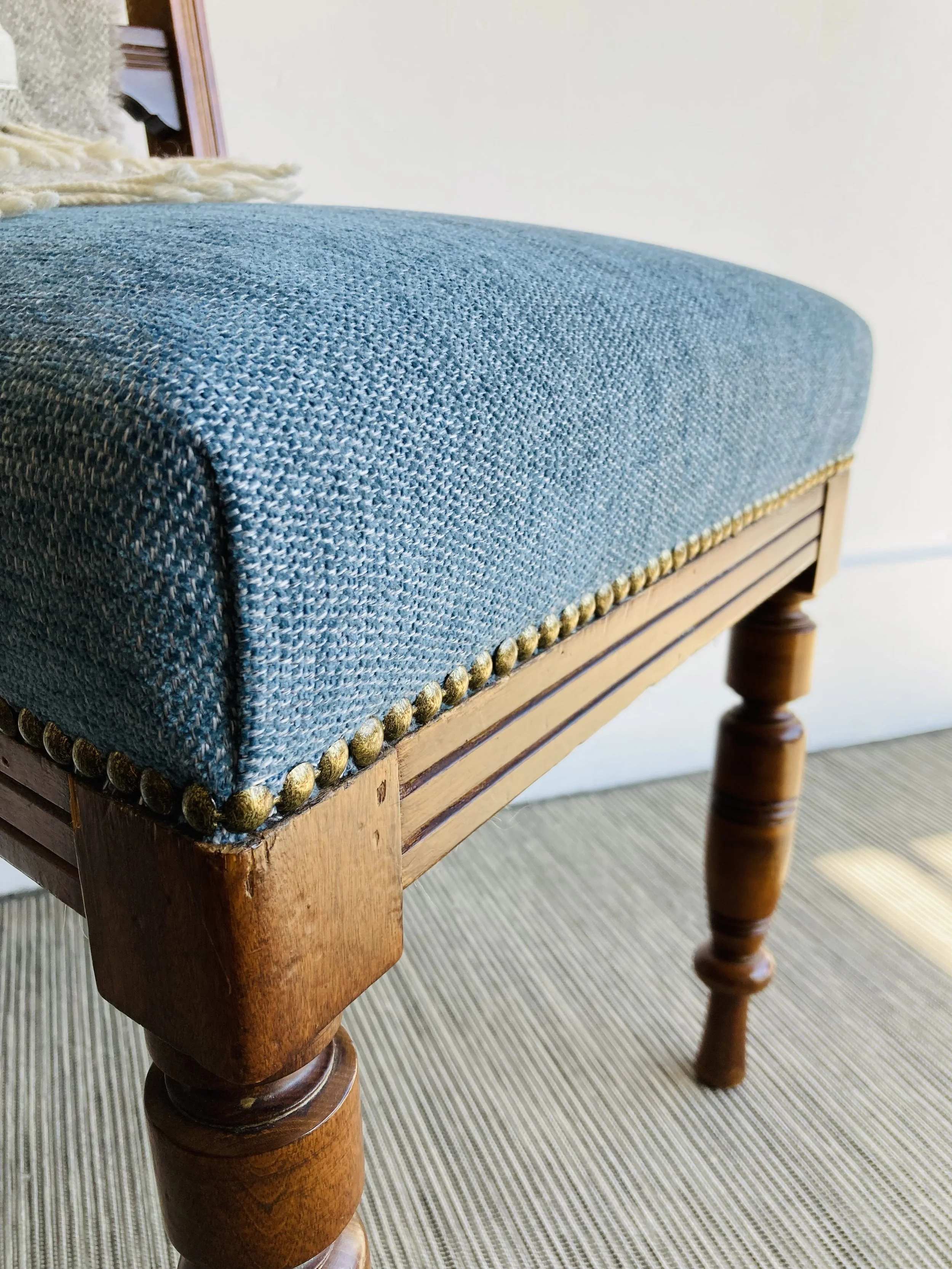 Close-up of a wooden table with a blue upholstered fabric top and decorative gold nailhead trim, showing part of a wooden leg and a beige carpeted floor.