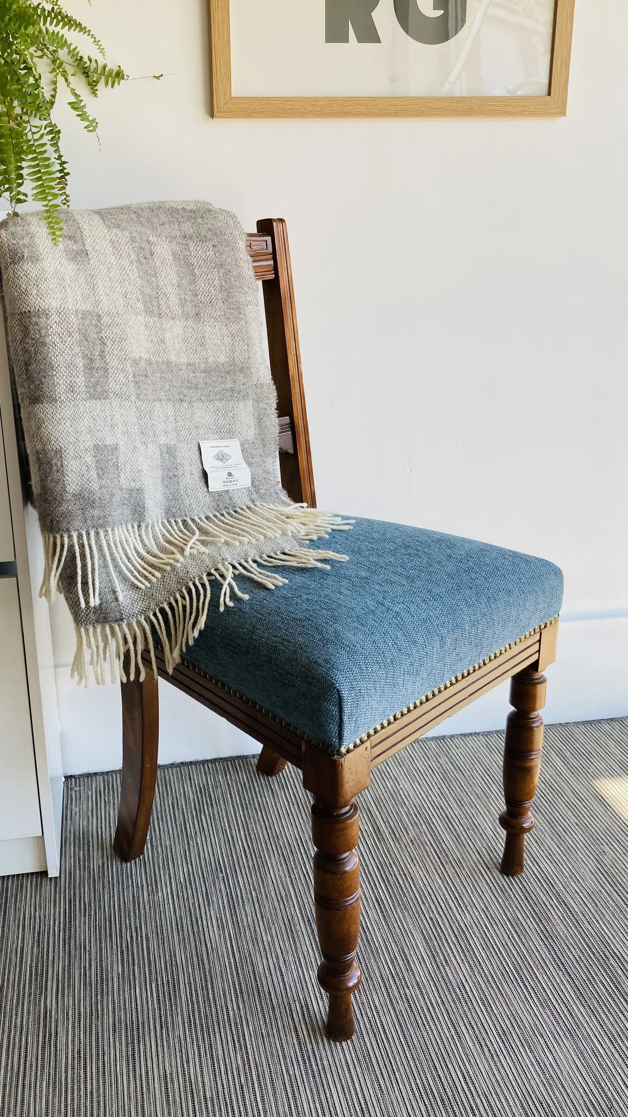 A wooden chair with blue upholstered seat and turned legs, placed on a striped gray and black carpet. A gray and cream woven throw blanket is draped over the backrest, and there's a framed piece of wall art hanging above the chair.