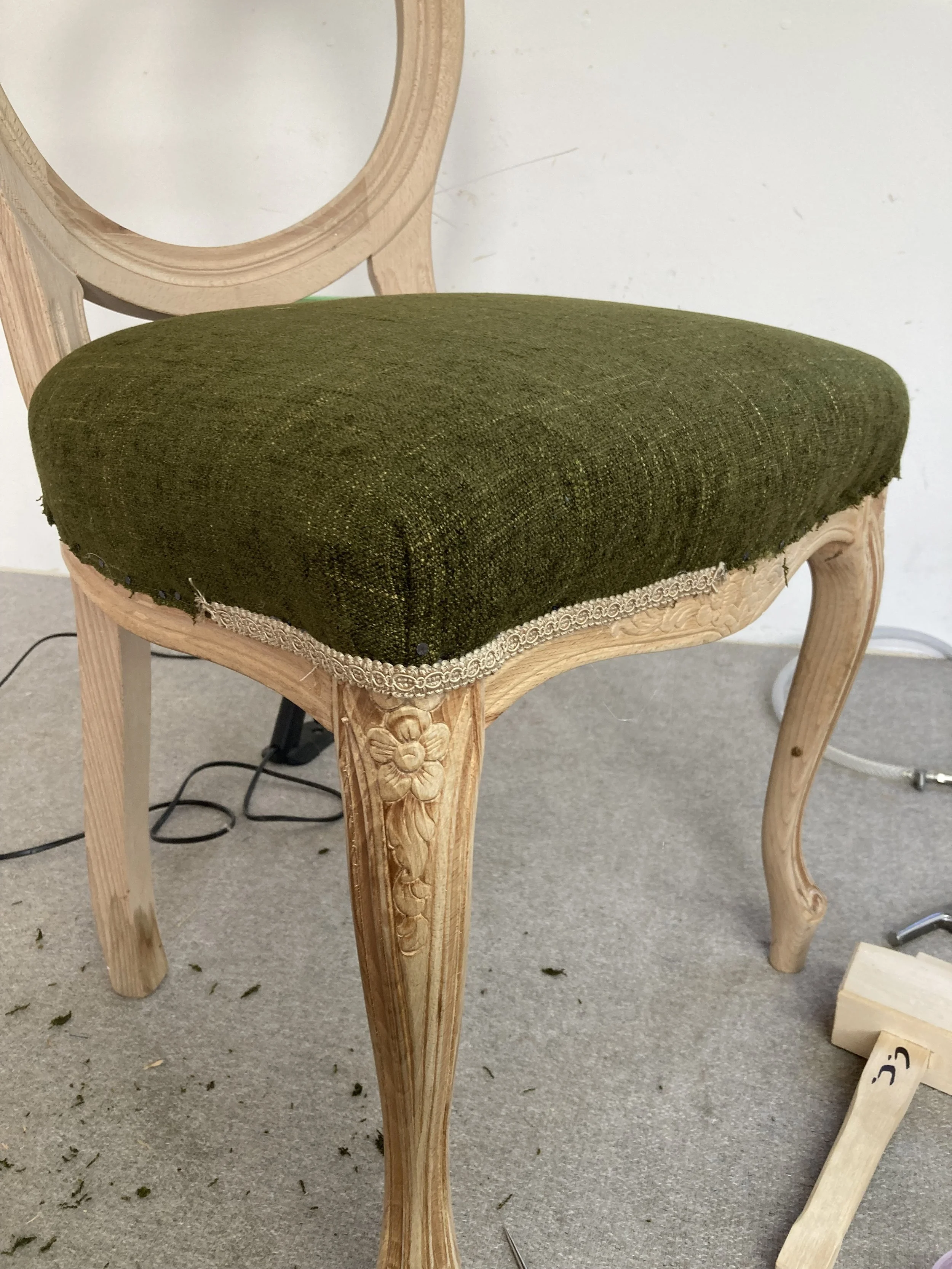 A wooden chair with a carved floral design on the front of its leg, a green upholstered seat, and a circular backrest frame, set on a carpeted floor with some dust and debris.