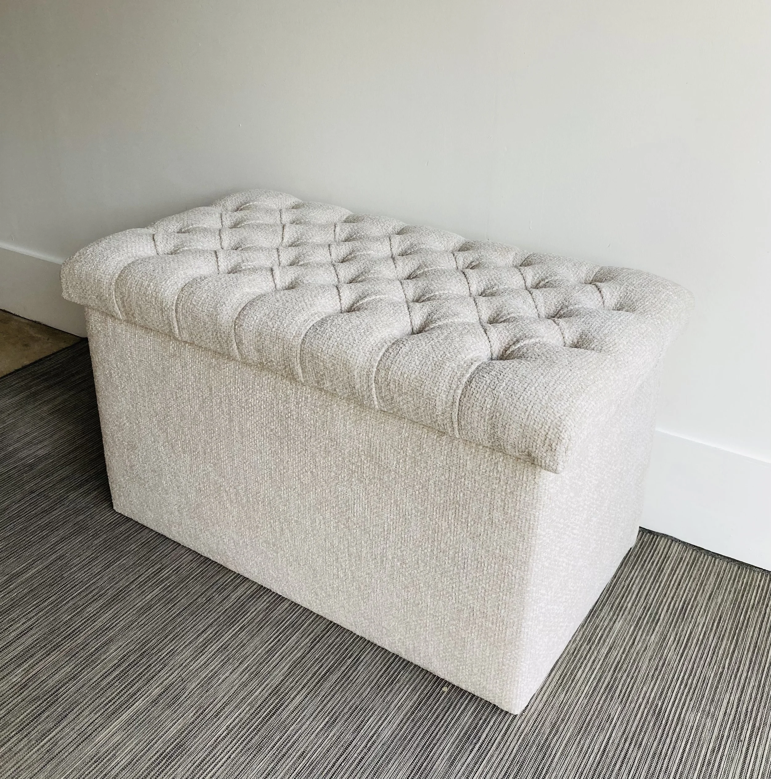 A beige tufted upholstered bench with a textured fabric sits against a light-colored wall and on a dark striped carpet.