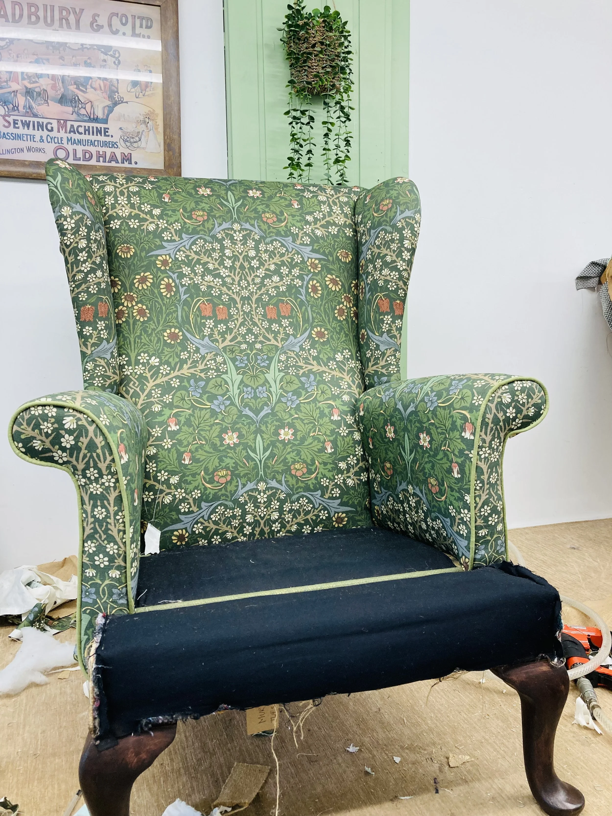 A sofa with a green floral patterned fabric backrest and armrests, and a black seat cushion, placed on a wooden floor. There is a poster and a hanging plant in the background, and some debris on the floor, indicating the sofa is being repaired or restored.
