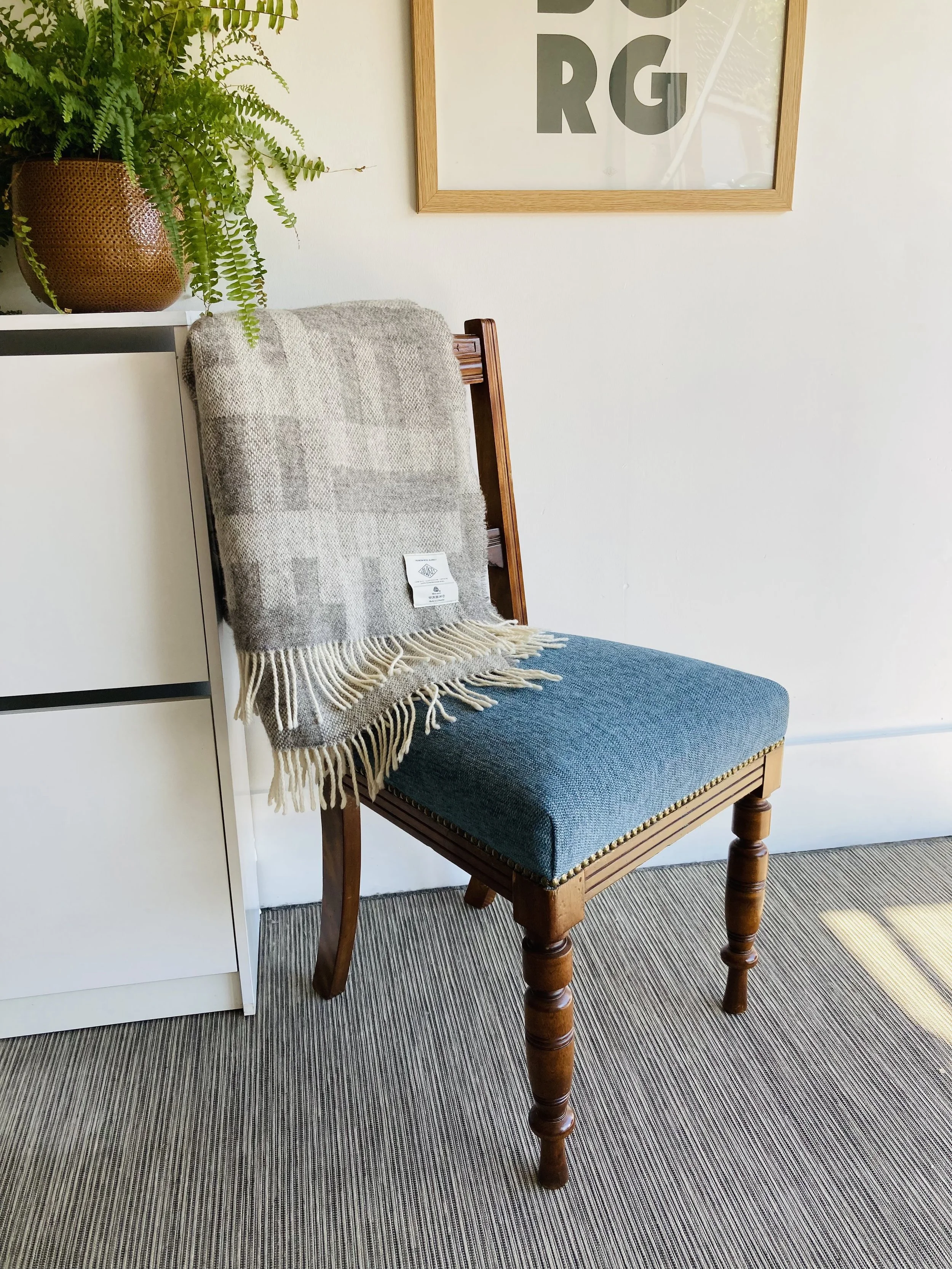 A wooden chair with a blue upholstered seat and a beige and gray plaid throw blanket draped over the backrest. To the left, there is a white shelving unit holding a potted fern plant. The wall behind the chair features a framed piece of art with bold black letters.