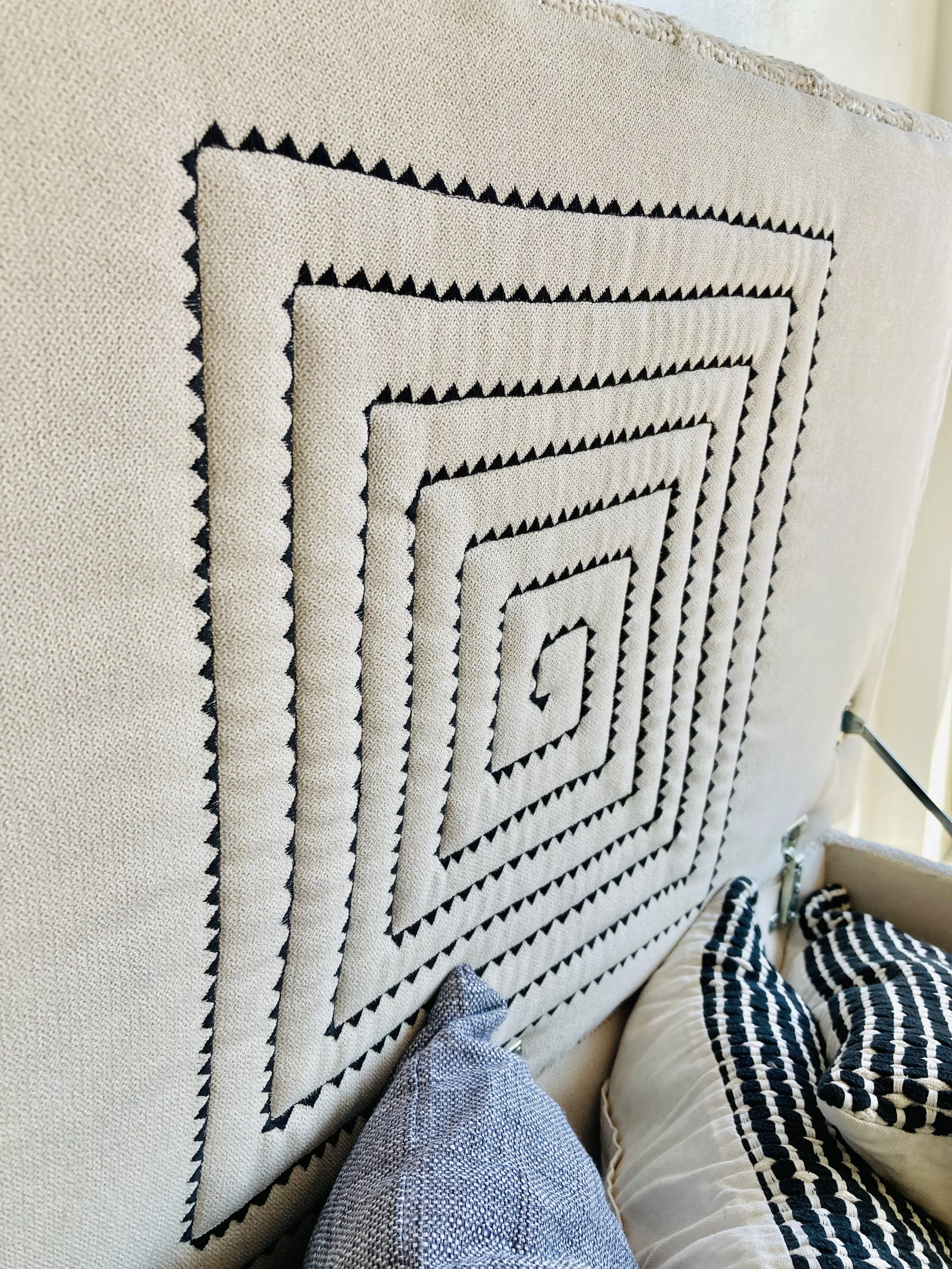 Close-up of a beige upholstered headboard with black embroidered geometric patterns, showing a person's legs in light gray textured pants and striped black and white pajamas, with part of a table leg visible.