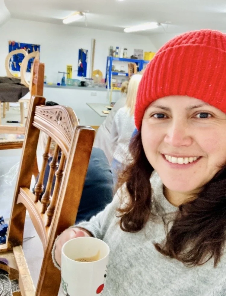 Woman in a red knit hat smiling, holding a mug, in a workshop or studio with chairs and shelves in the background.