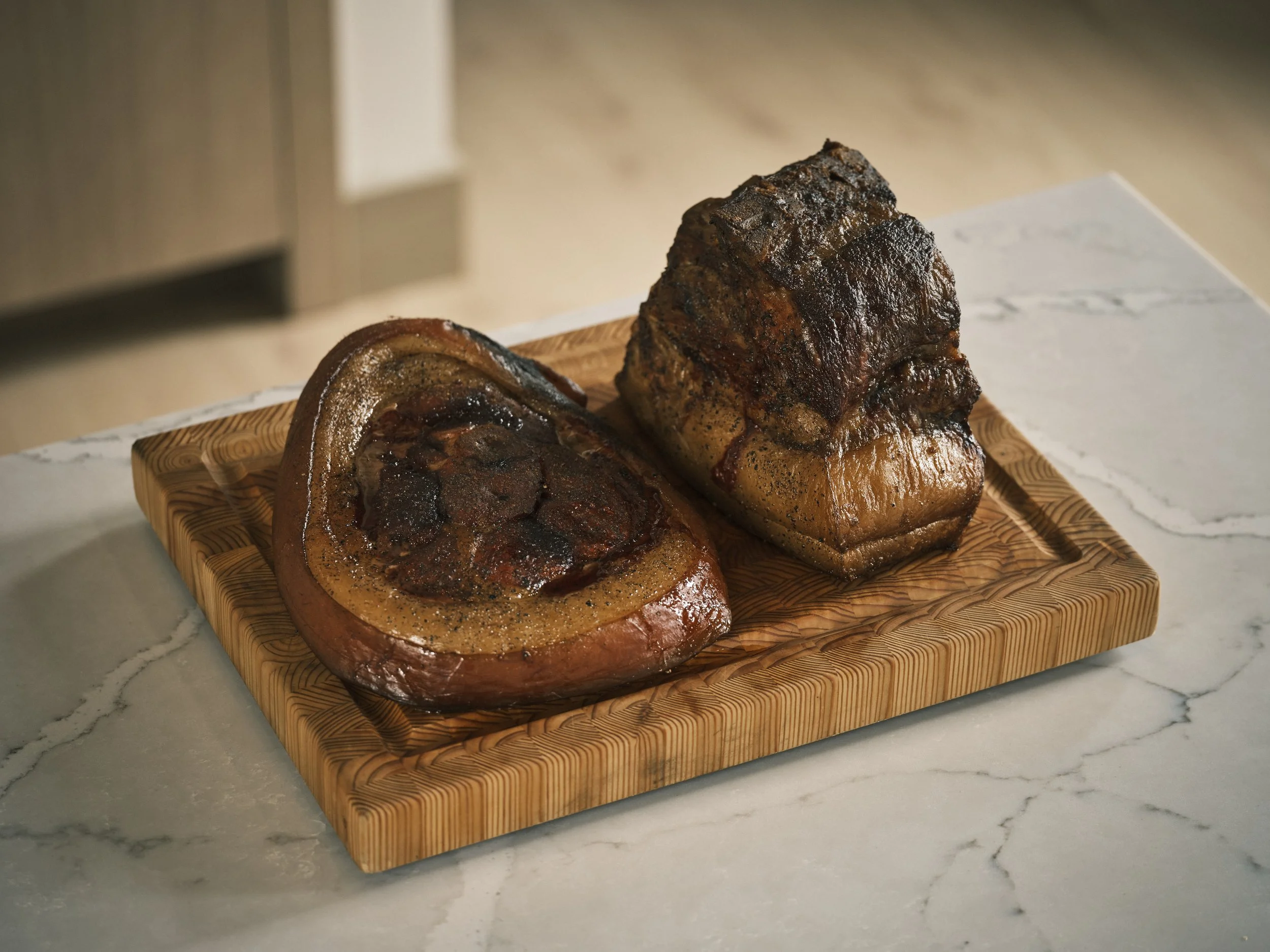 Two cuts of cooked beef on a wooden cutting board, placed on a white marble surface.